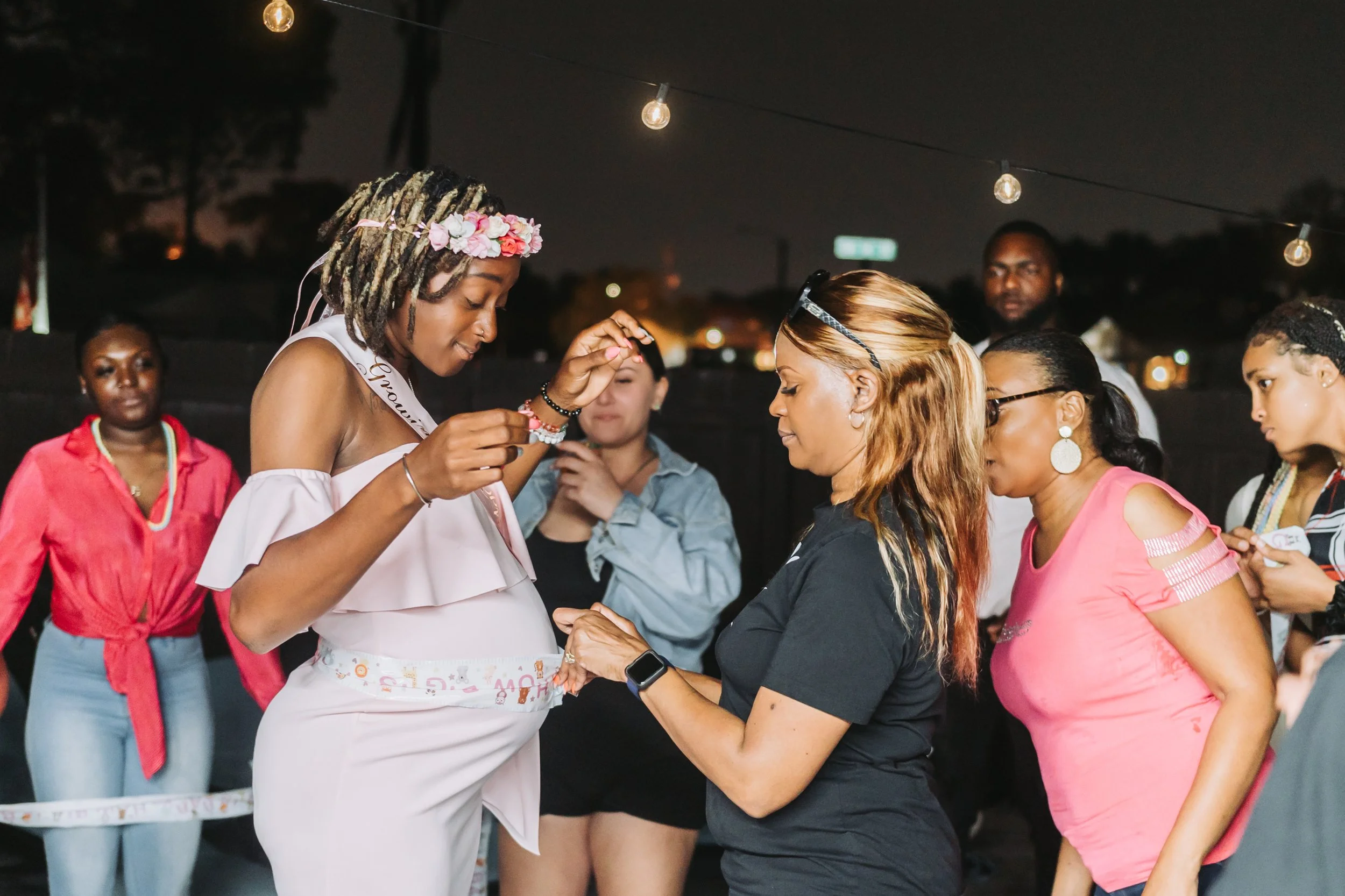 A woman with dreadlocks and a flower crown is placing a necklace on another woman with light brown hair at an outdoor event at night. Several women and a man are observing, all dressed casually, and there are string lights hanging overhead.