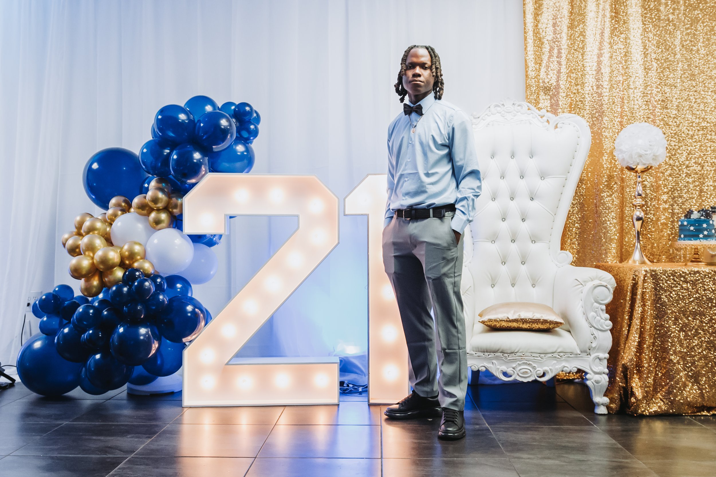 Young man in formal attire standing beside decorative balloons, a large illuminated number two, and a white ornate chair with gold accents at a celebration or party.
