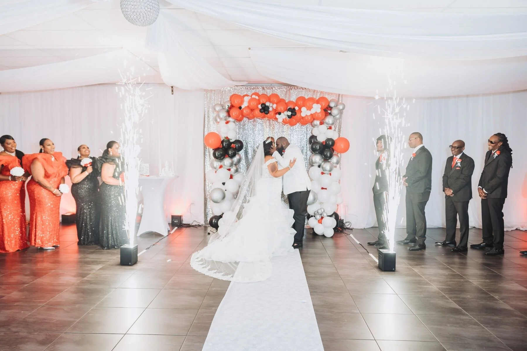 A wedding ceremony with a bride and groom dancing in the center, surrounded by their wedding party and guests. The room is decorated with balloons, streamers, and sparklers.