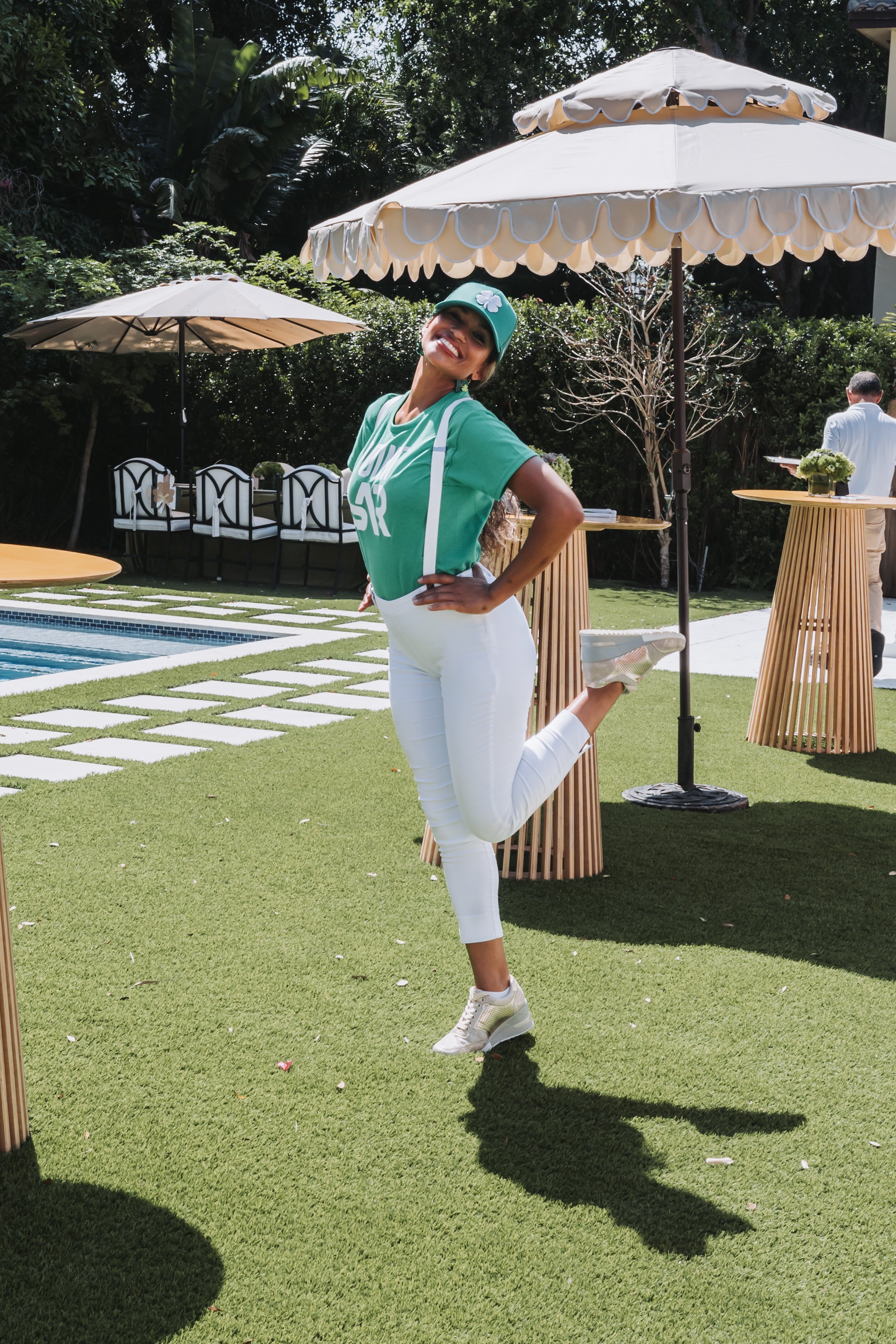 A woman in a green shirt, white pants, and sneakers is smiling and posing with one leg bent behind her on a lawn. She is wearing a green cap and standing near patio tables with large beige umbrellas, surrounded by greenery and outdoor furniture.