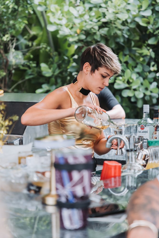 A woman with short hair wearing a gold top and black choker pouring a drink from a bottle at an outdoor bar with bottles and bar tools surrounded by greenery.