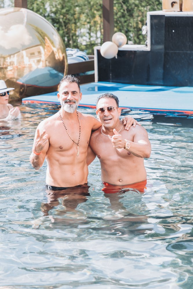 Two men enjoying a swim in a pool, smiling and making gestures at the camera. One is wearing sunglasses, another a beaded necklace, and both are shirtless. A woman is visible in the background, and decorative spheres are seen near the poolside.