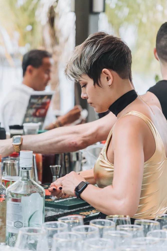 A woman with short, two-toned hair and a black choker, wearing a gold shiny top, is preparing drinks at a bar. There are various bottles and glasses on the counter, with other people in the background.