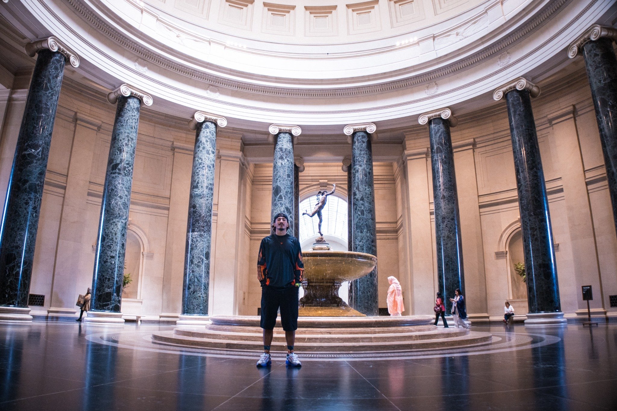 A person standing inside a grand, circular interior with tall marble columns, a high domed ceiling, and a statue of a woman holding a torch in the background.