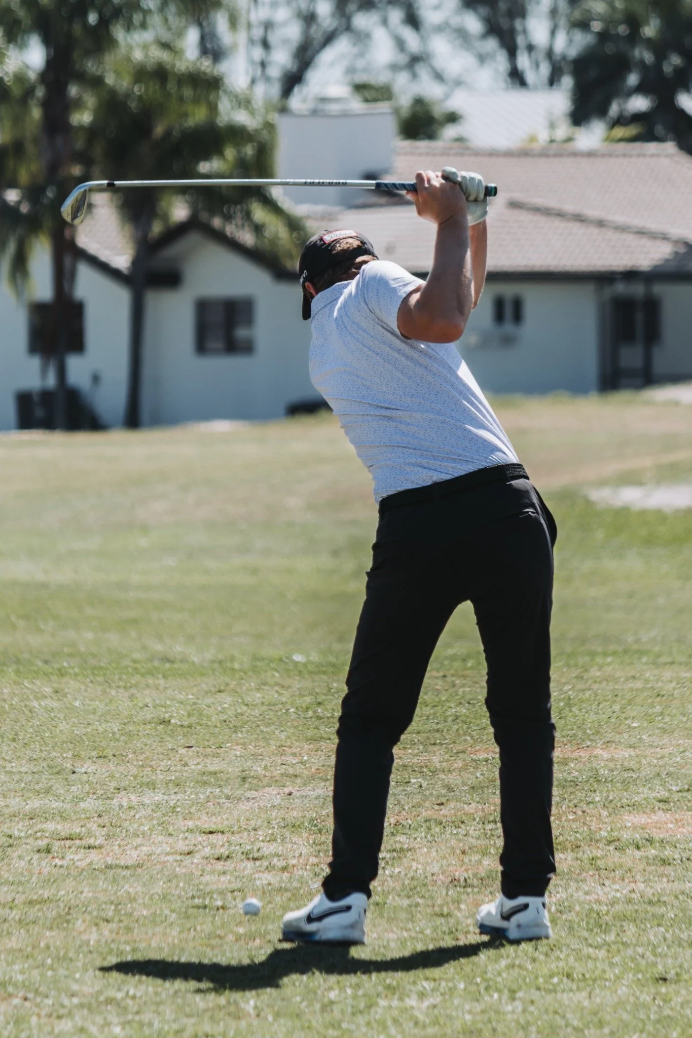 A man wearing a white golf shirt, black pants, and a black cap swings a golf club on a golf course with houses in the background.