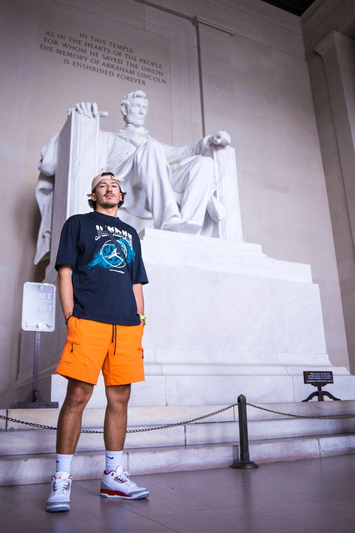A young man stands in front of the Lincoln Memorial in Washington D.C., wearing a black t-shirt, orange shorts, white sneakers, and a beige cap, with the large seated statue of Abraham Lincoln in the background.