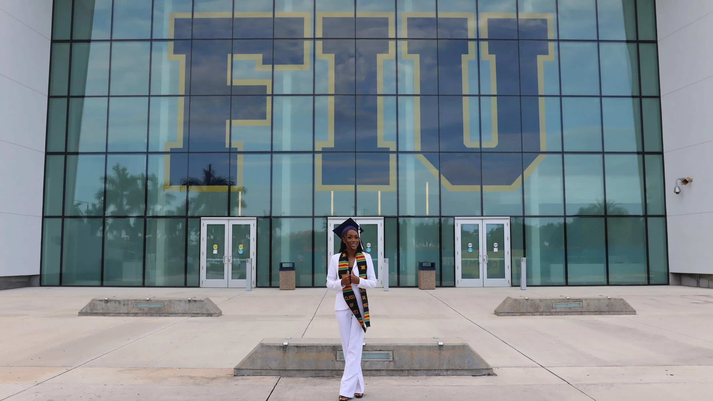 A woman dressed in a white graduation gown and cap, wearing a multicolored graduation stole, standing in front of a large glass building with the year 2023 displayed prominently on the windows, smiling with her hands clasped in front of her.