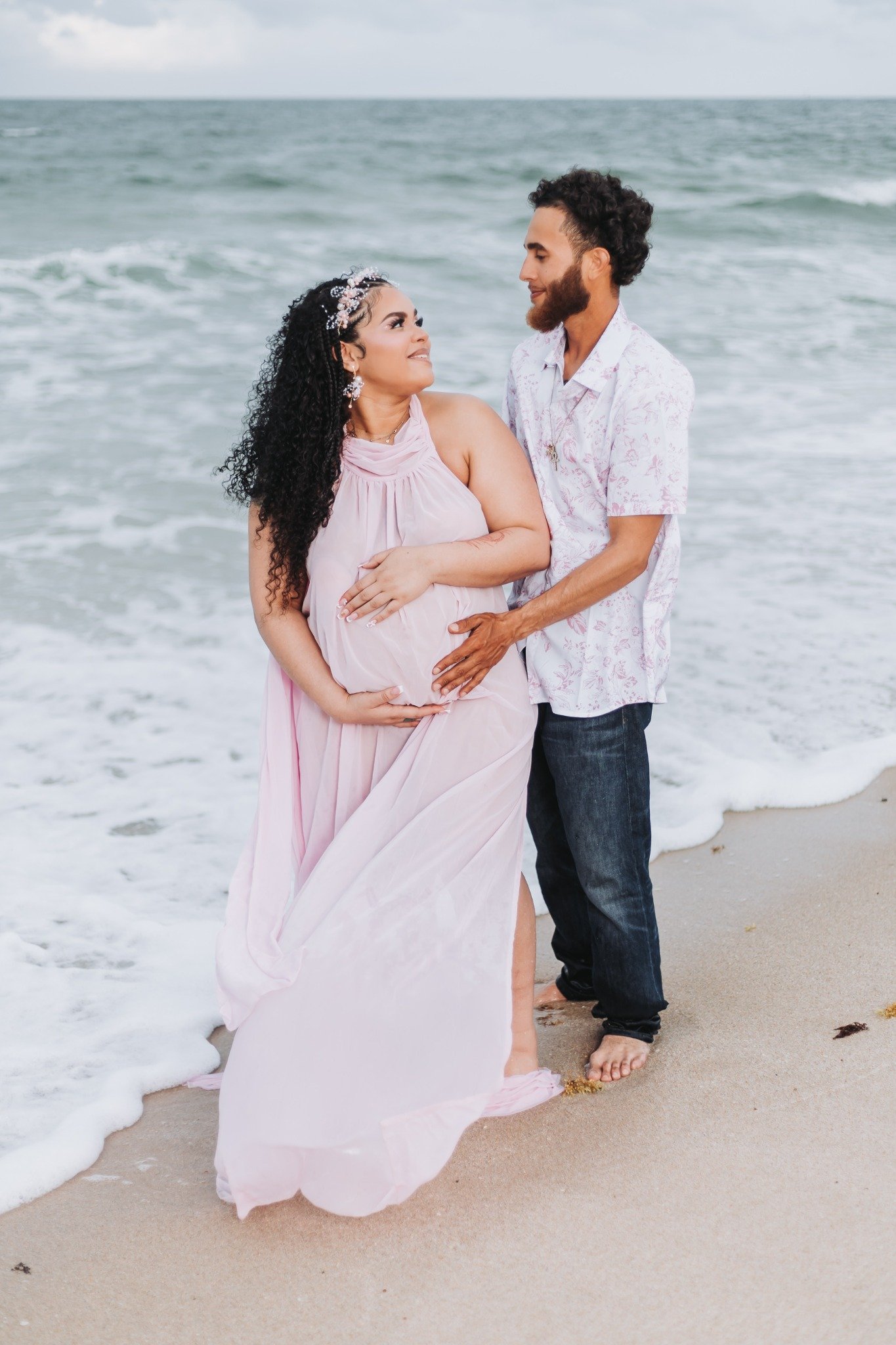 A couple standing on the beach near the water, with the woman wearing a pink dress and the man in a floral shirt and jeans, both barefoot on the sand.