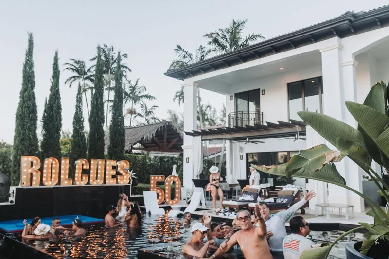 People enjoying a pool party in front of a modern white house with large glass doors, surrounded by tall palm trees and greenery, with a marquee sign spelling 'ROLGUES' and a large number '50' candle.