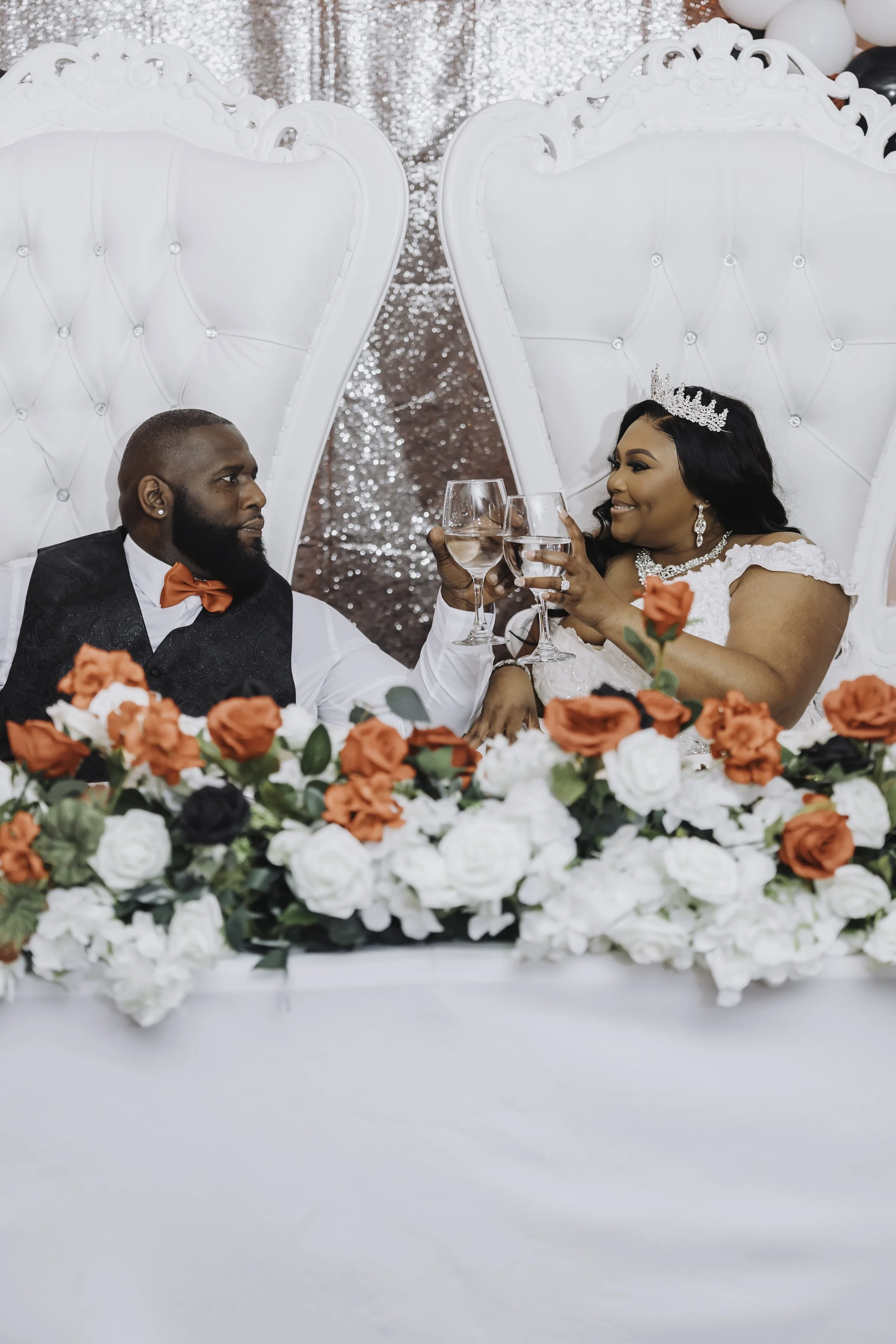 A bride and groom celebrating at their wedding reception, raising glasses in a toast. The bride is wearing a white dress with lace details, a tiara, and jewelry. The groom is in a black suit with a red bow tie. They are sitting at a table decorated w