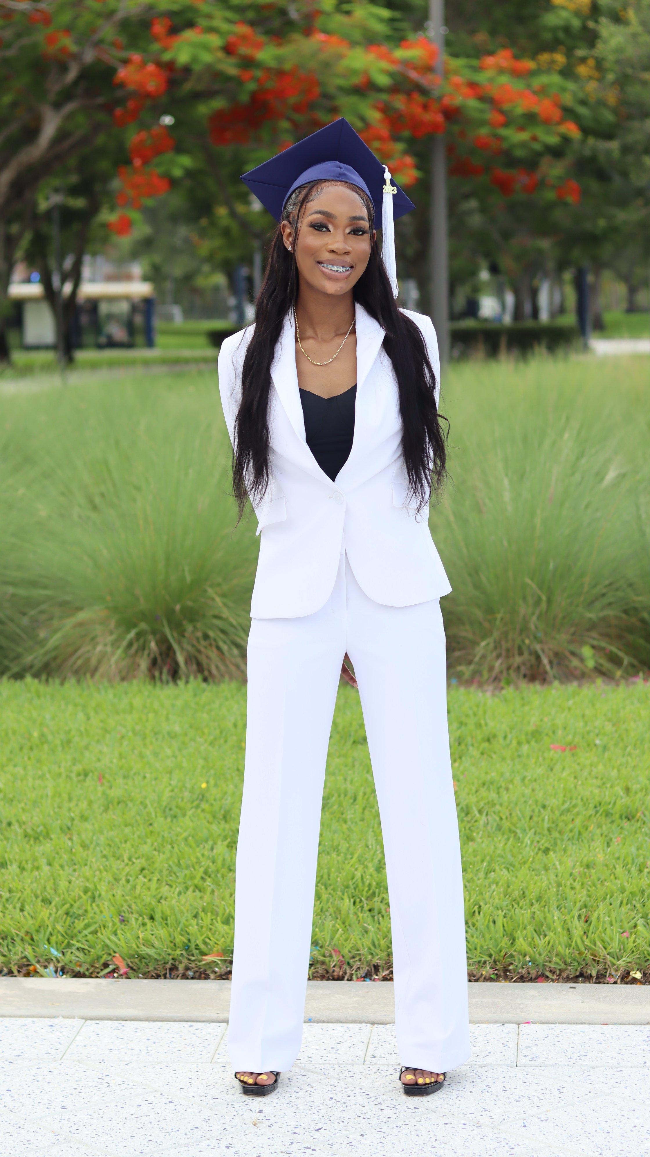 A young woman in a white suit and black top standing outdoors with red and green trees in the background. She is wearing a graduation cap with a white tassel and has long black hair. She is smiling and wearing open-toed heels.