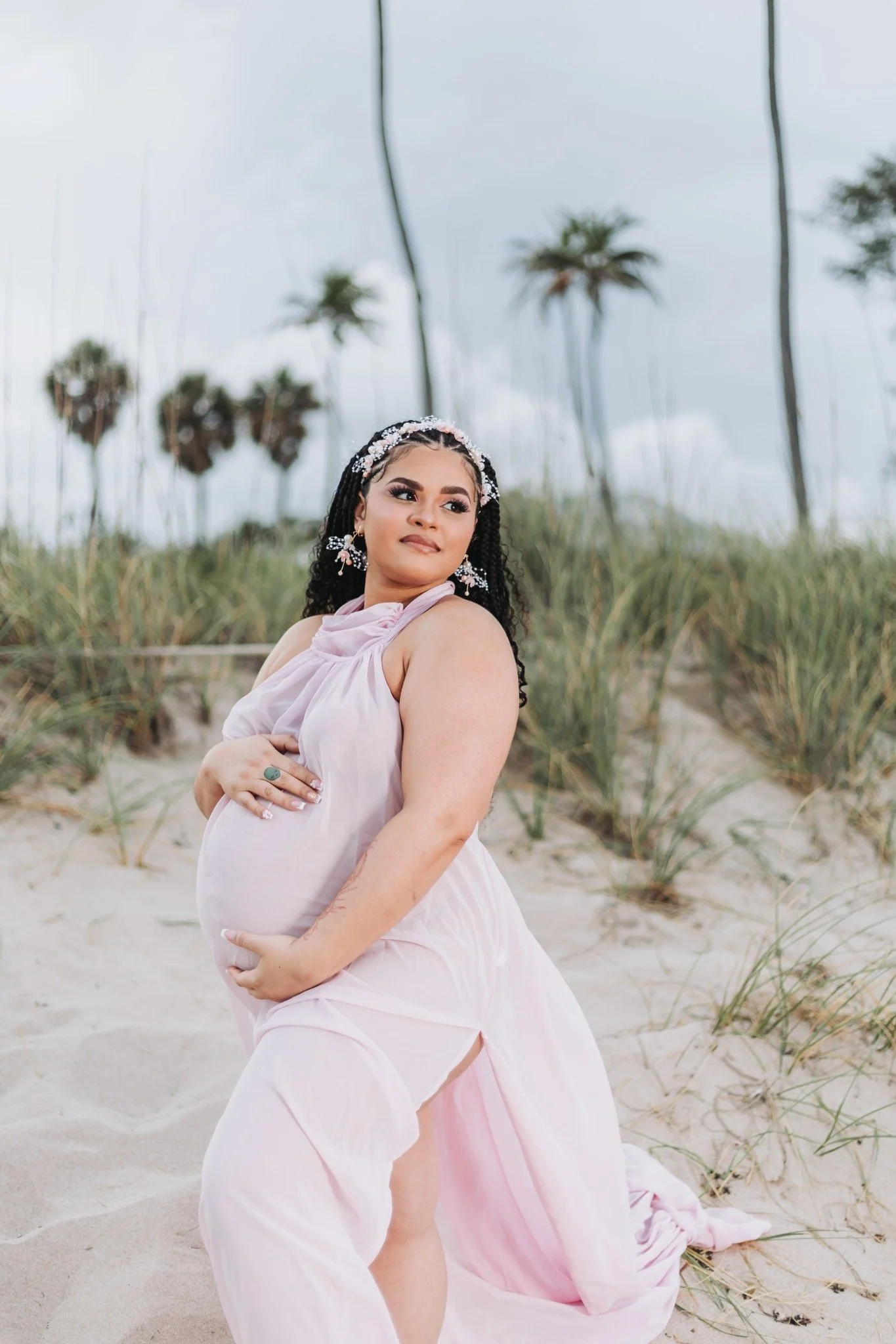 A pregnant woman in a flowing pink dress standing on a beach with sand and grass, with palm trees in the background, looking to the side with a gentle smile.