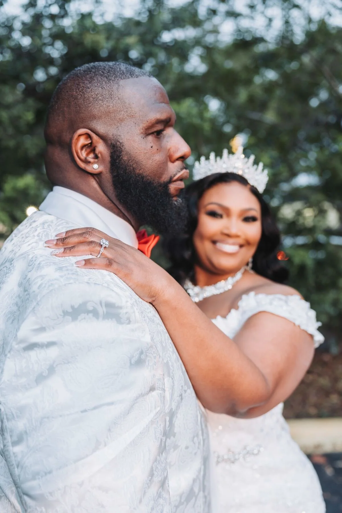 A groom in a white tuxedo with a red bow tie and a bride in a white wedding gown with a tiara, smiling in an outdoor setting with trees in the background.
