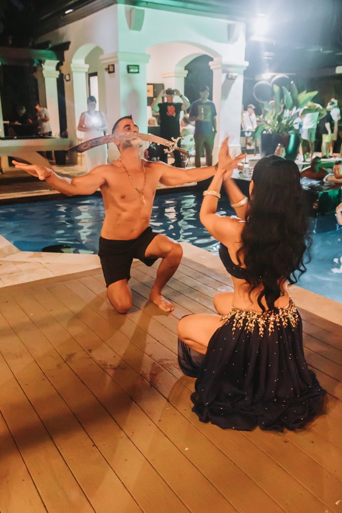A man performing a snake dance with a large snake near a swimming pool at a nighttime party. A woman in a black dress watches, surrounded by other people.