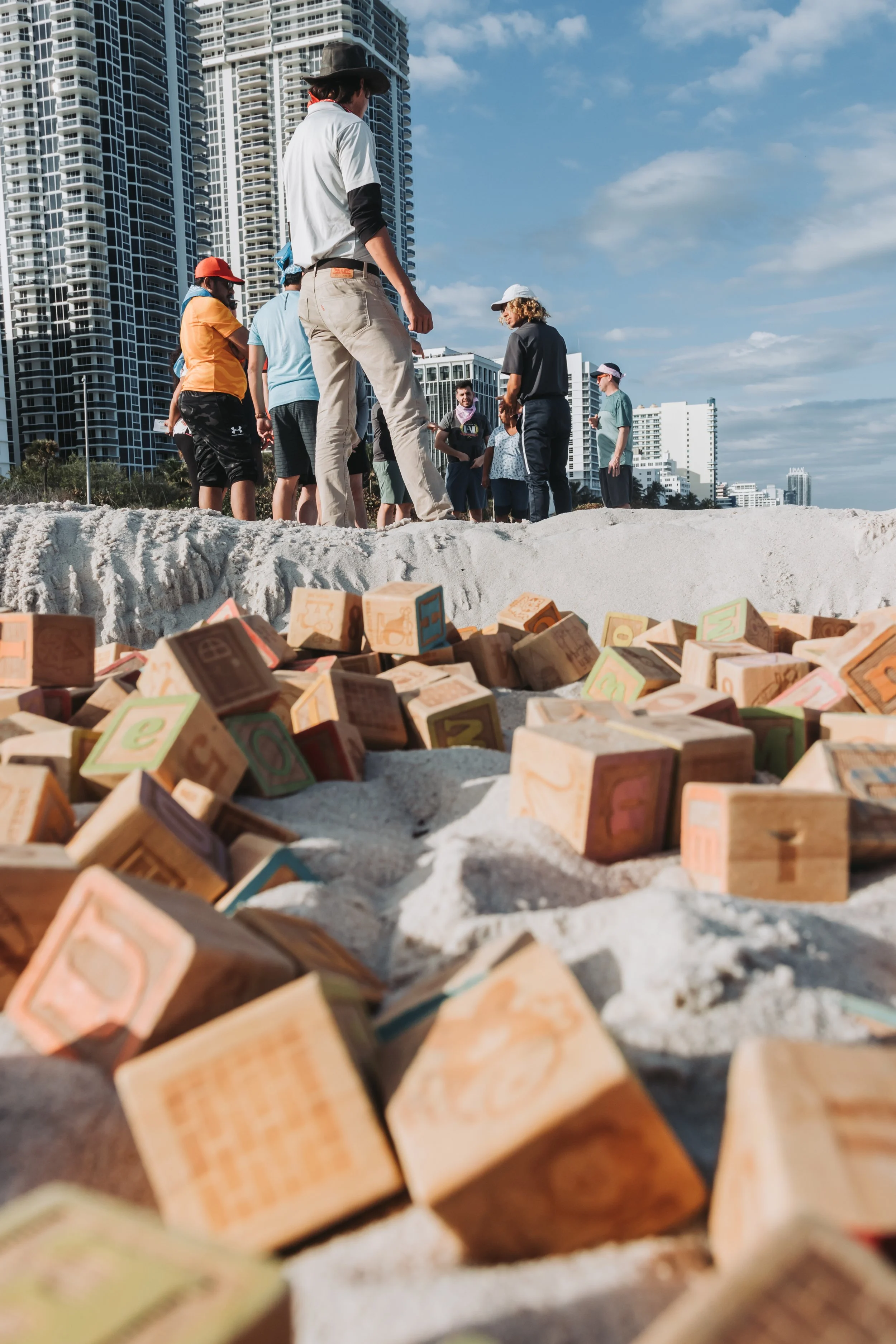 People gather around on a sandy beach with high-rise buildings in the background, some wearing hats and casual clothing, with wooden blocks scattered on the sand in the foreground.