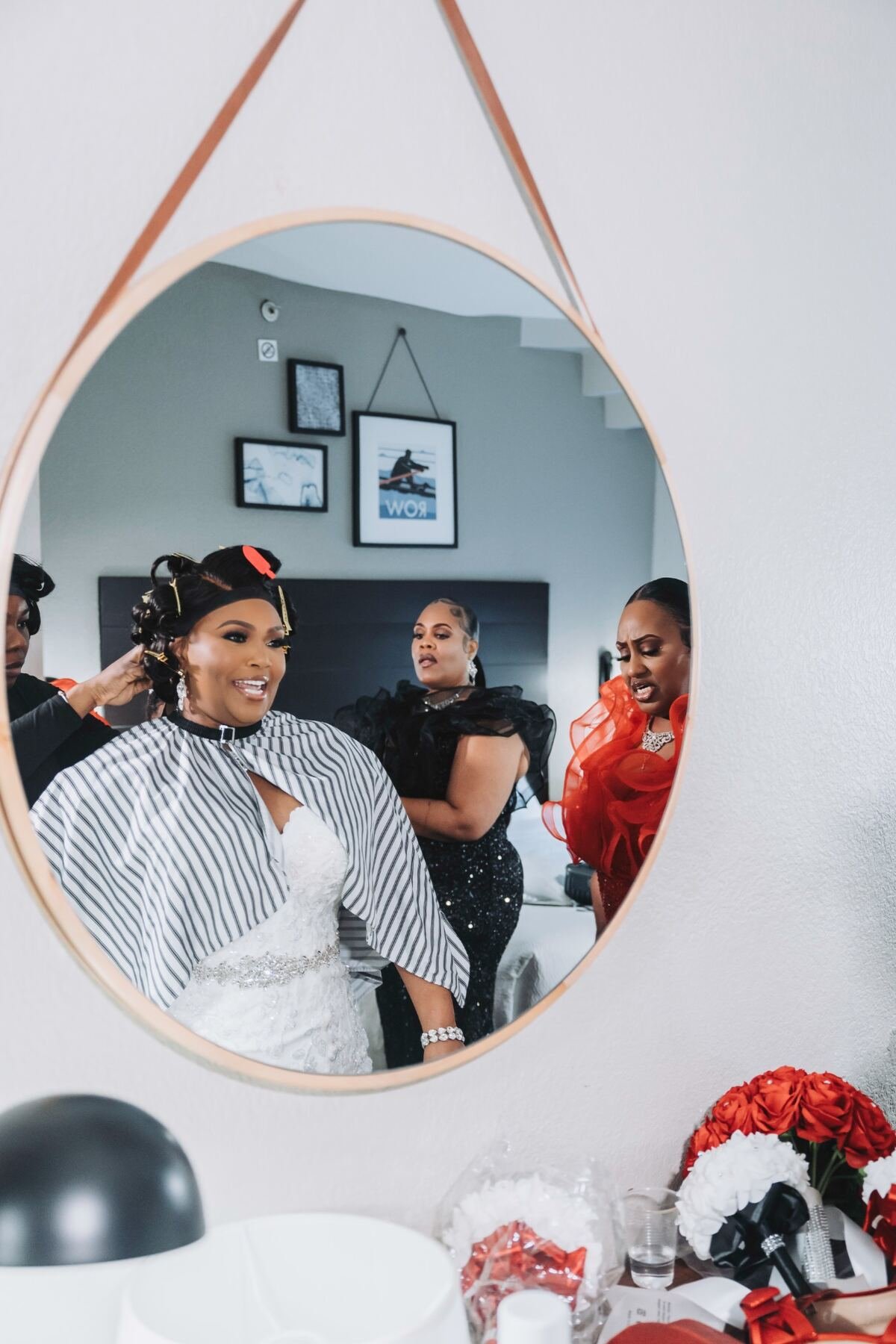 Three women getting ready, reflected in an oval mirror. One woman in a white wedding dress is smiling, while another woman in a red dress looks concerned and a third woman in black stands behind them with crossed arms.