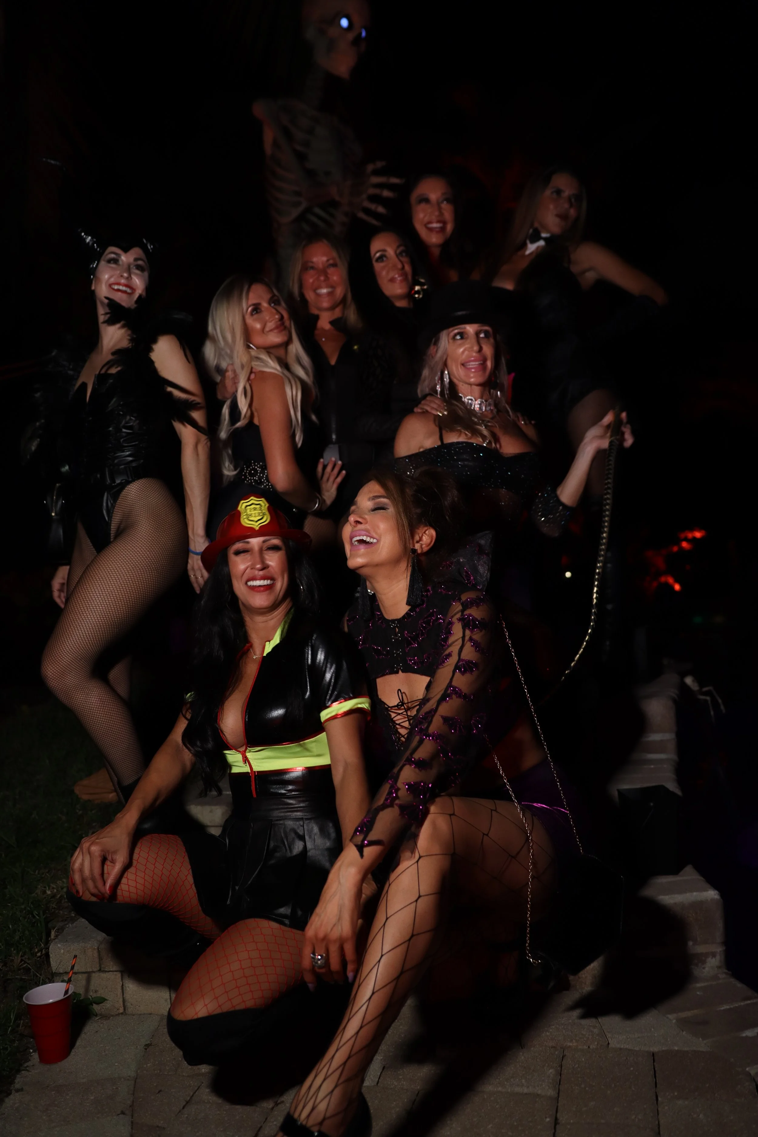 Group of women in Halloween costumes, including a devil, a skeleton, a witch, and a police officer, gathered outdoors at night celebrating. Some are smiling and posing for the camera.