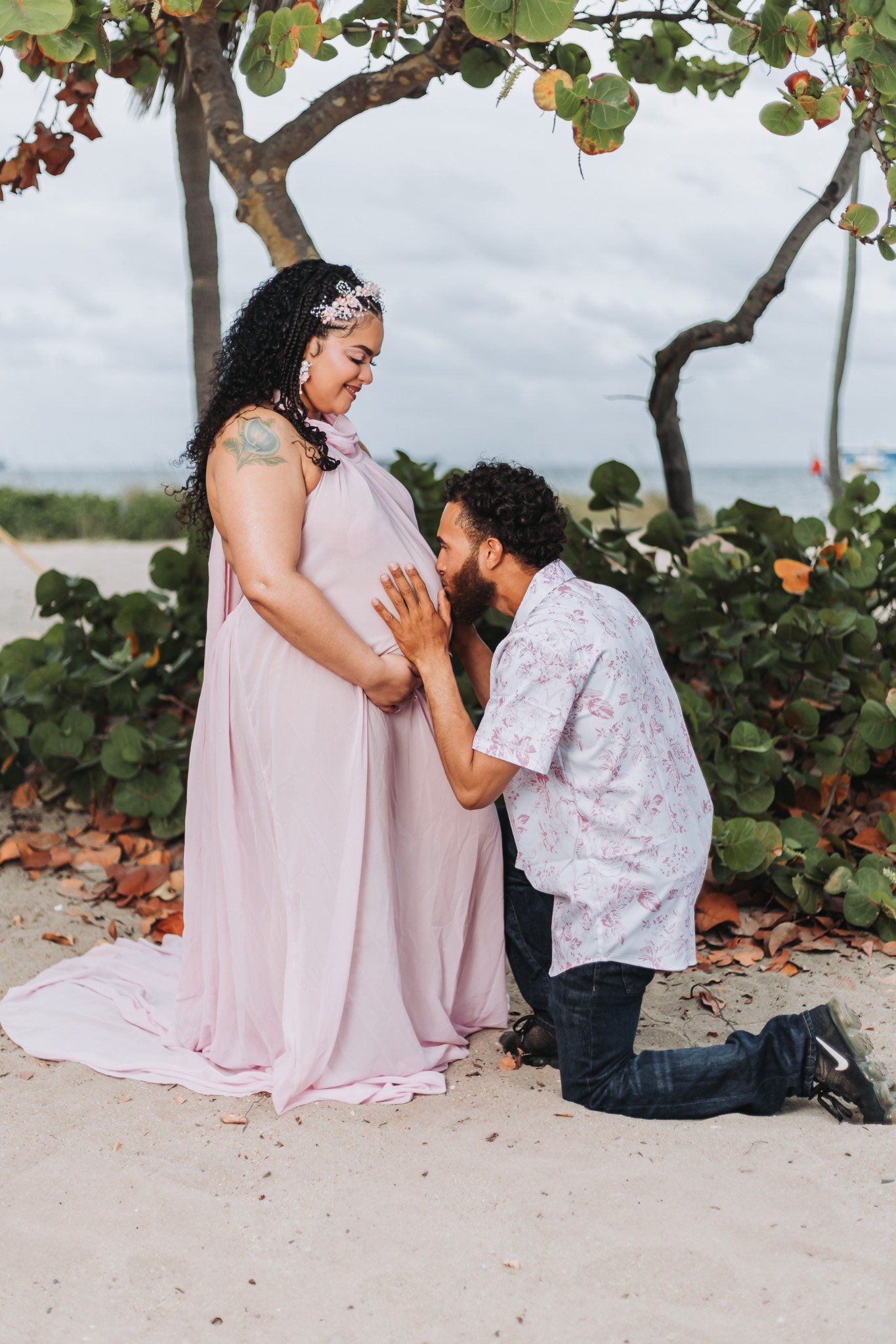 A pregnant woman standing on a beach under a tree, with a man kneeling before her and kissing her belly, in a romantic scene near the ocean.