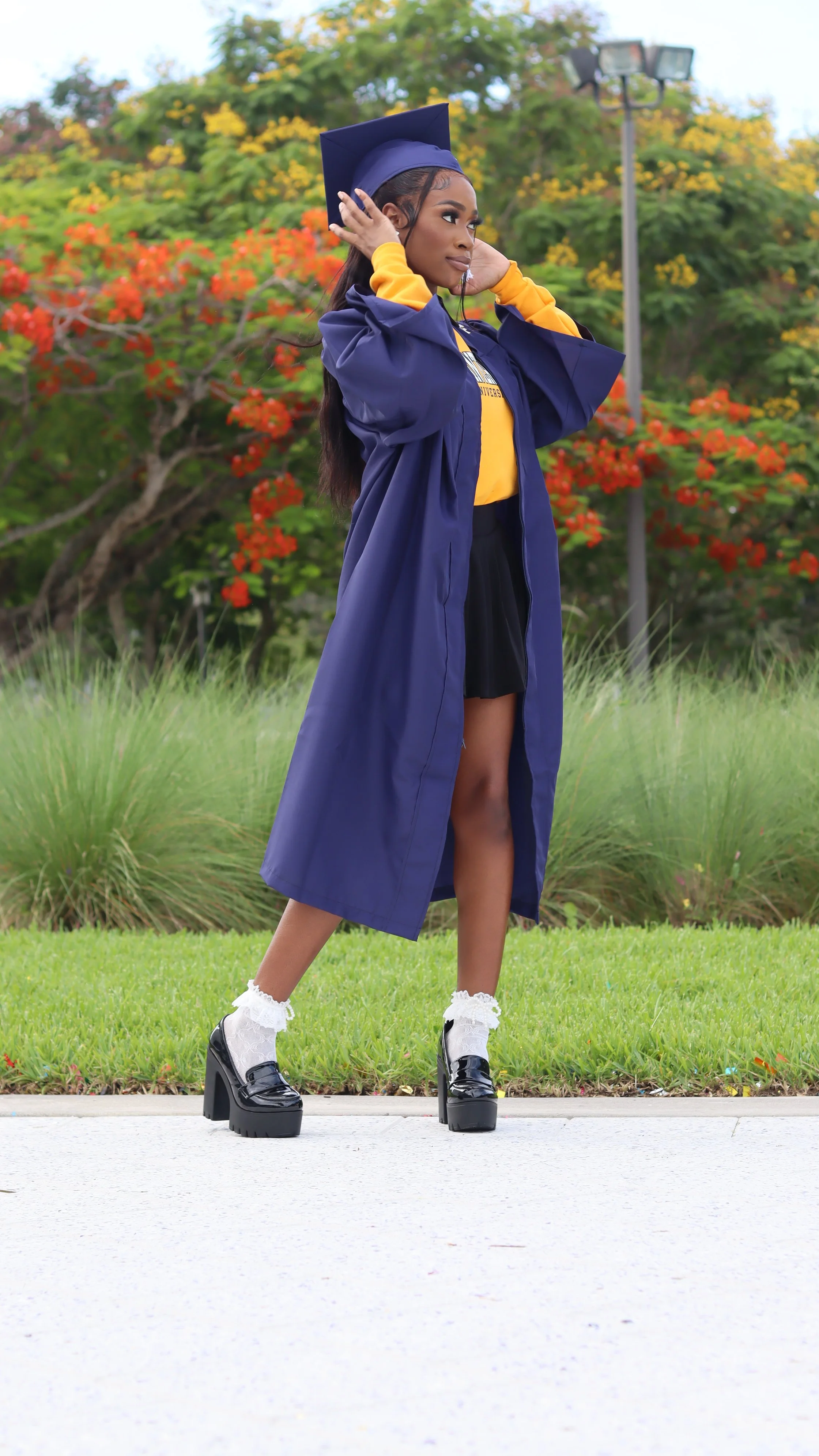 Young woman in a graduation cap and gown, standing outdoors on grass with colorful trees and a lamp post in the background, adjusting her cap.