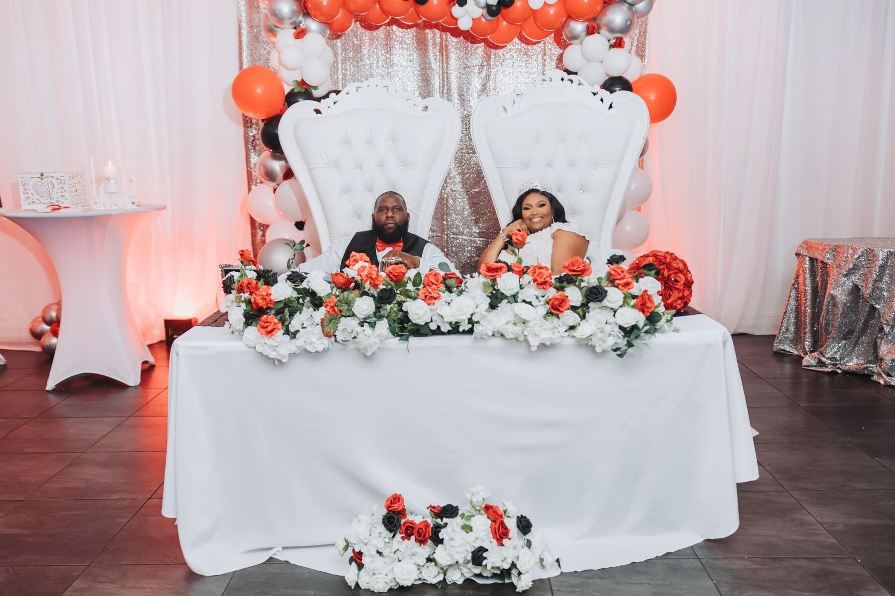 A couple seated at a decorated table for a celebration, possibly a wedding, with large white high-backed chairs, floral arrangements of roses in black, white, red, and peach, and a balloon arch with black, white, red, and silver balloons behind them.