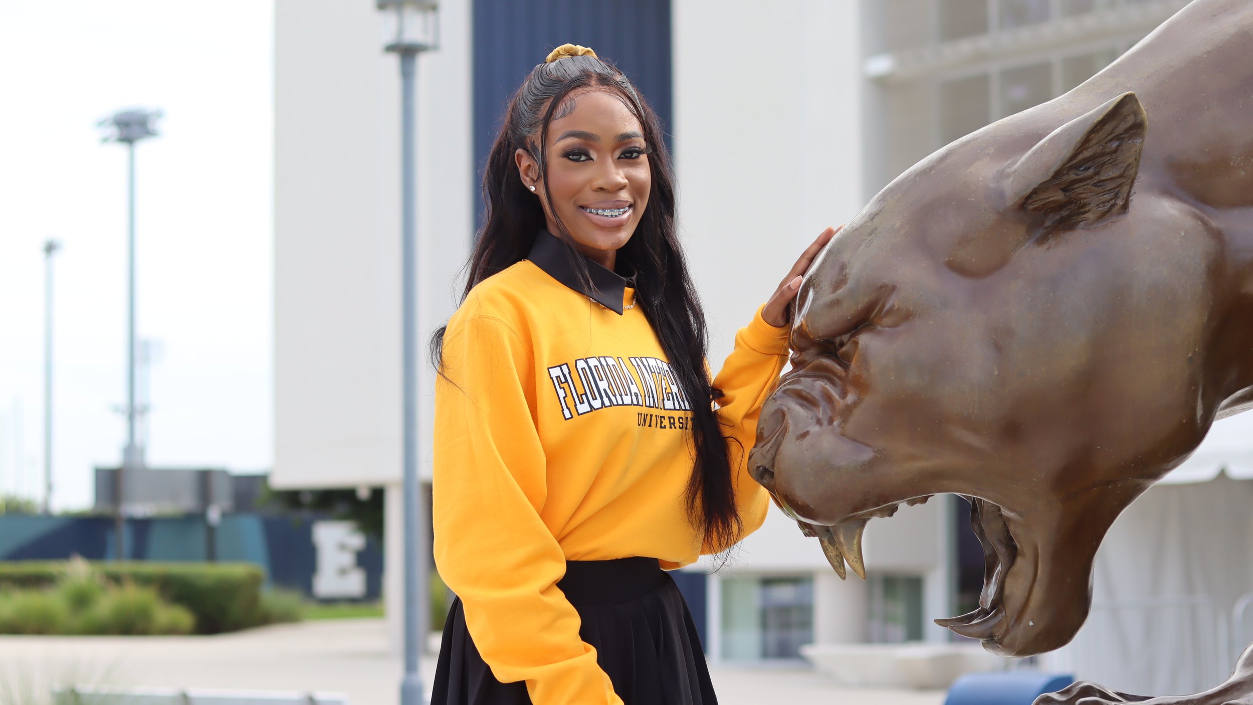 Young woman smiling and posing with a bronze horse head sculpture outdoors, wearing a yellow Florida A&M University sweatshirt.