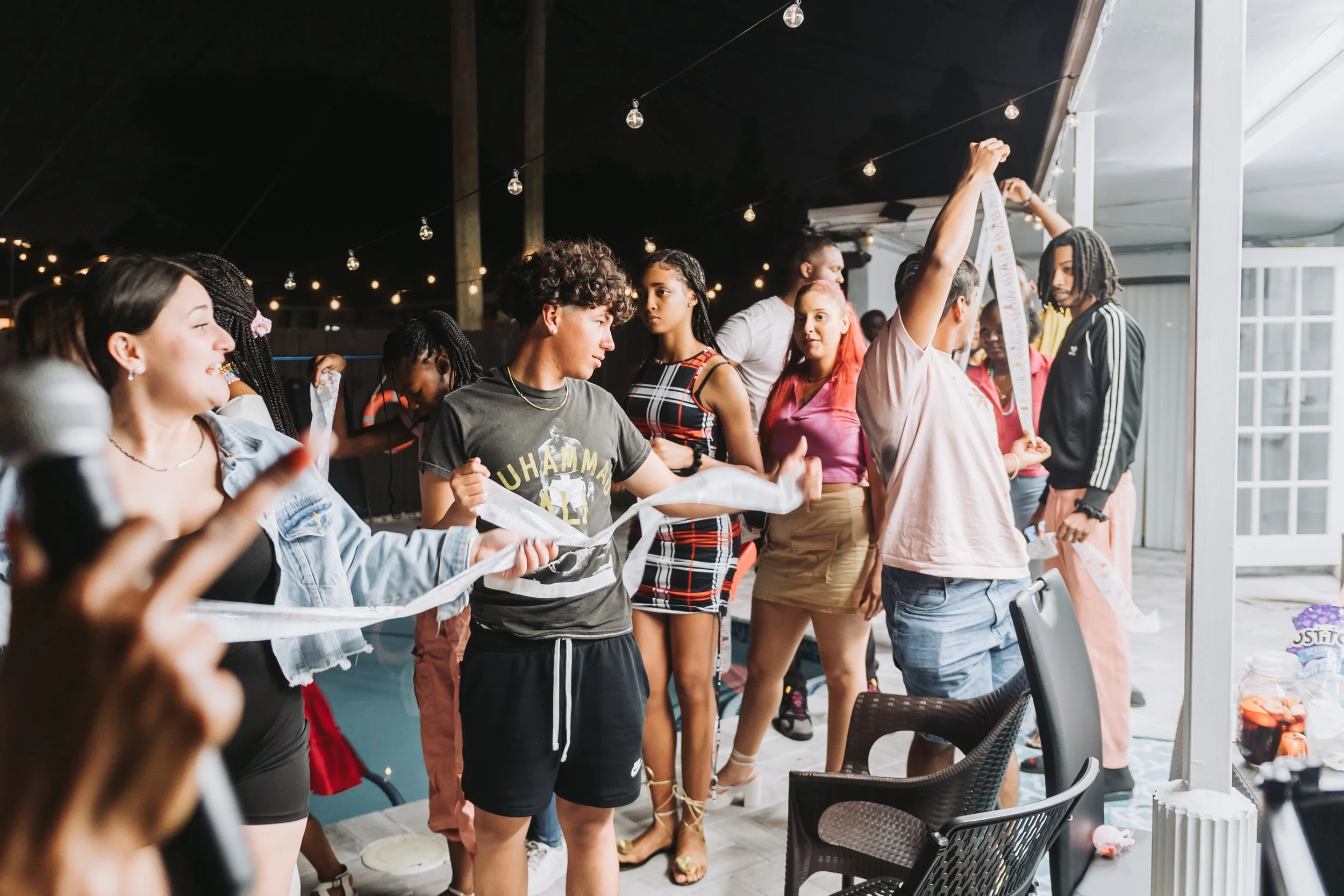 A group of diverse young people standing in a line, holding a long ribbon, at night under string lights, in an outdoor or semi-outdoor setting.