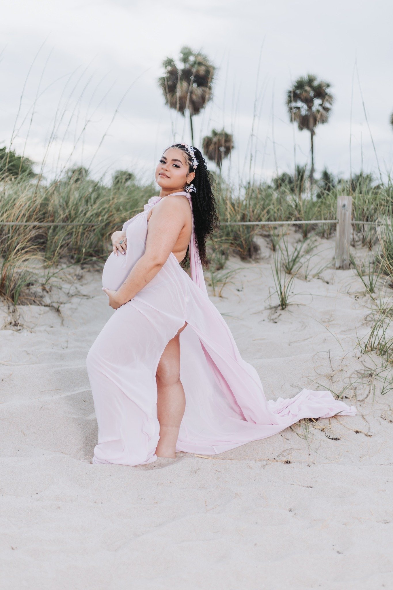 A pregnant woman in a flowing pink dress poses on a sandy beach with tall grass and palm trees in the background.