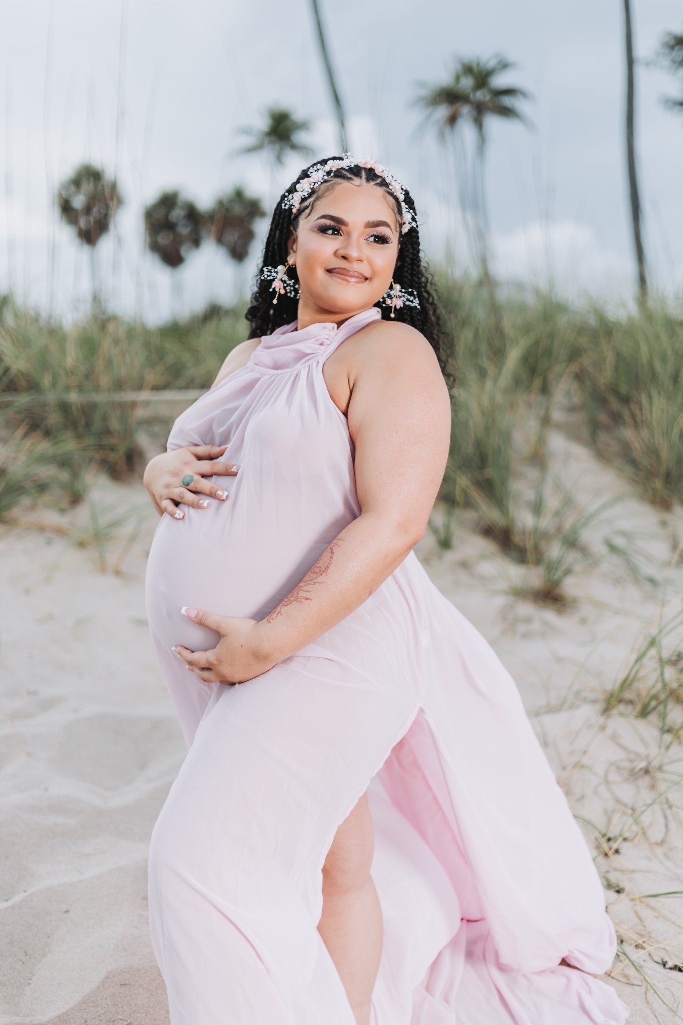 Pregnant woman in a pink dress standing on a beach with palm trees in the background.