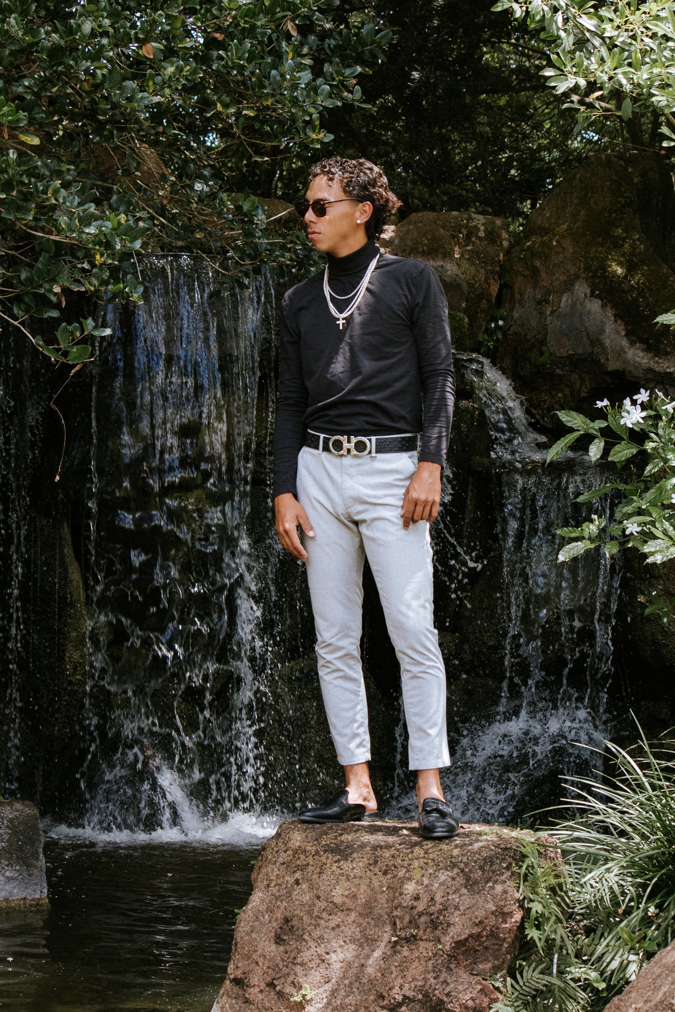 A man wearing sunglasses, a black turtleneck, layered necklaces, light-colored pants, and black loafers standing on a large rock in front of a small waterfall surrounded by green foliage.