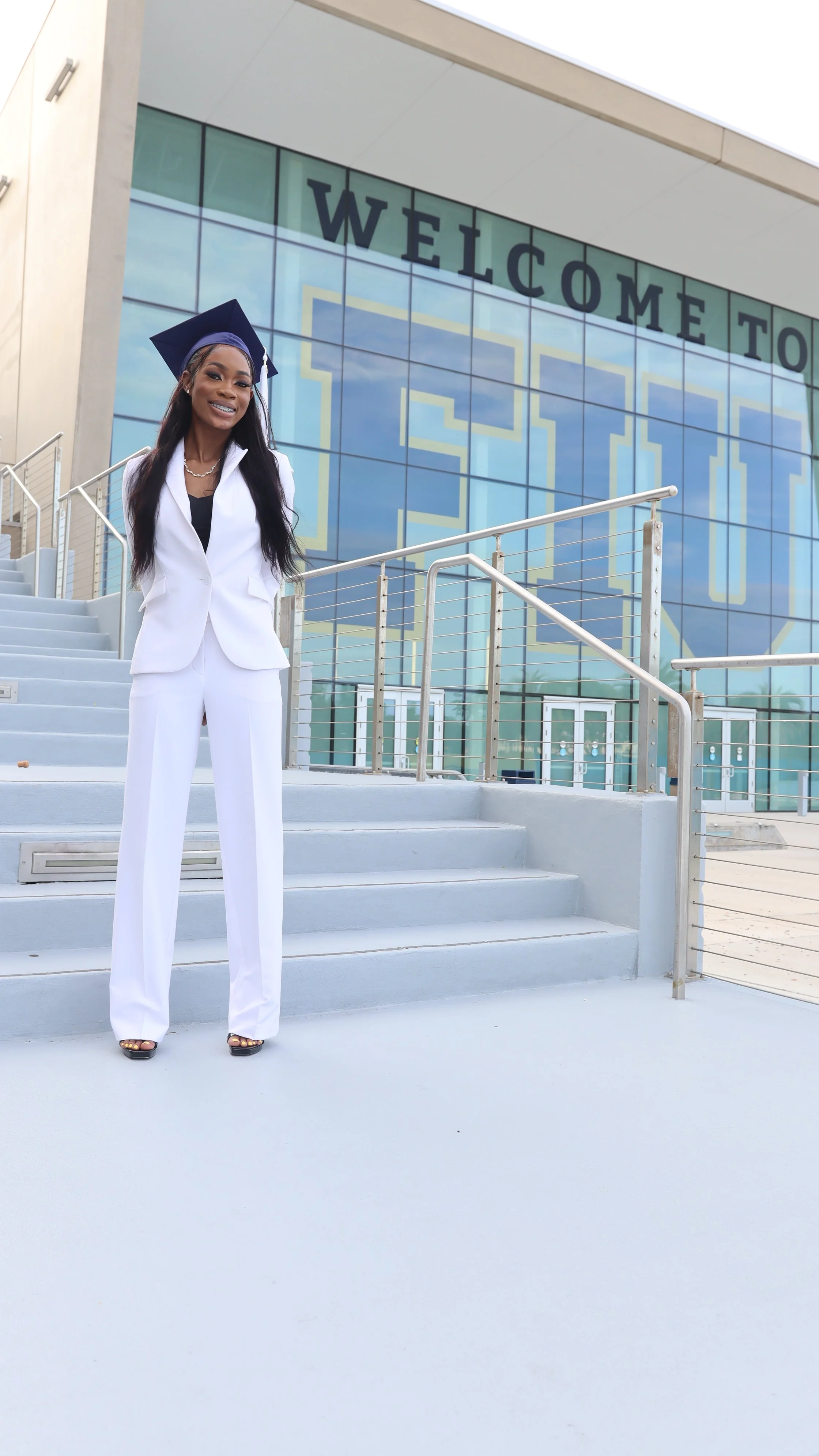 A woman in a white suit and graduation cap stands on steps outside a modern building with a large glass facade that displays the words "Welcome to".