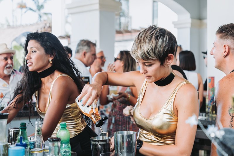 Two women in gold tops and black choker necklaces preparing drinks at a crowded party.