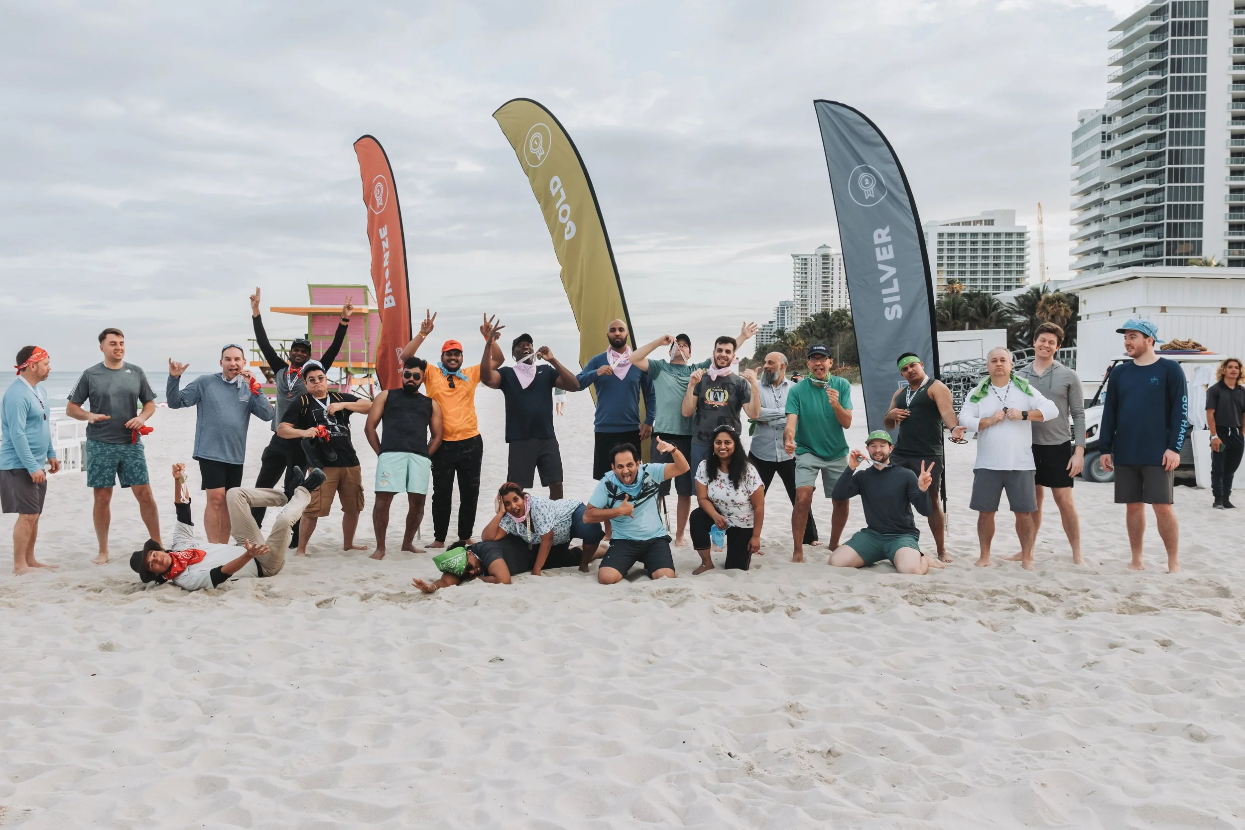Group of people on a beach posing for a photo with flags and high-rise buildings in the background, some making peace signs and smiling.