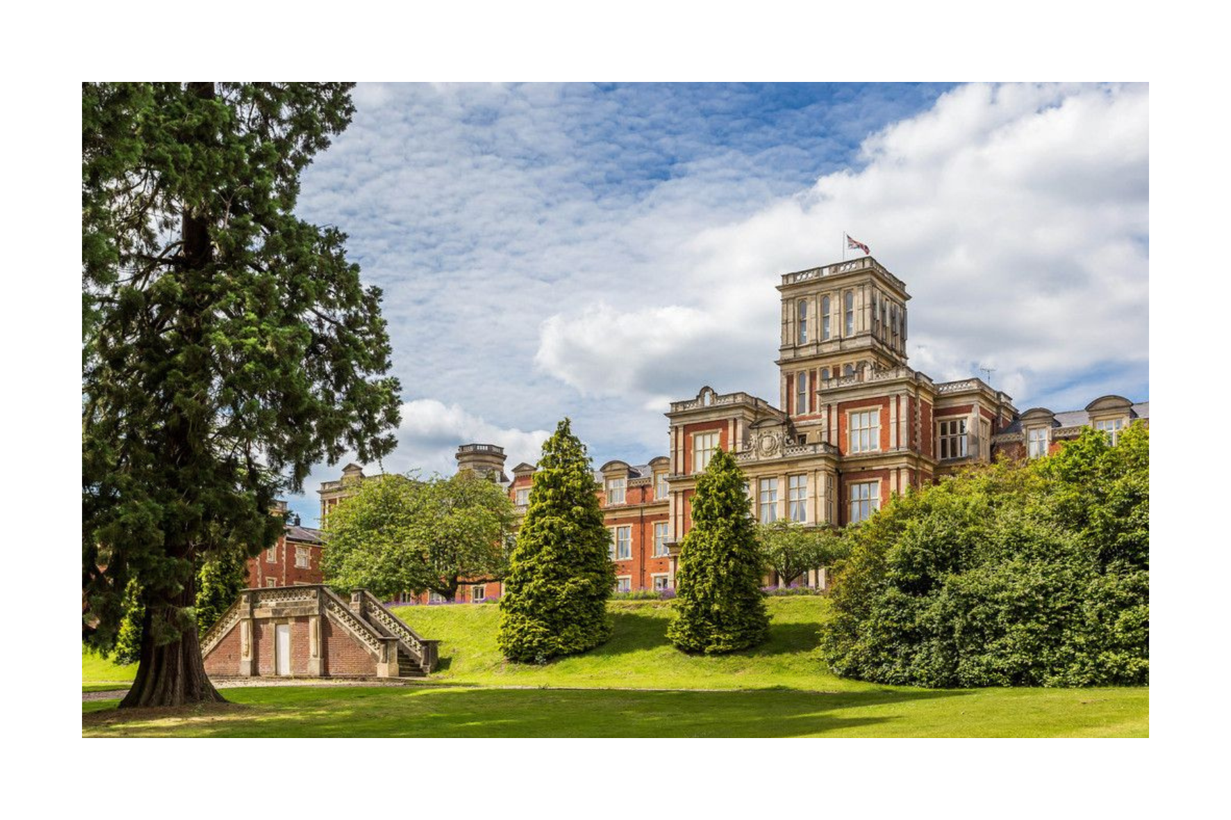 A historic red brick building with a tower, surrounded by green trees and a lawn, under a partly cloudy sky.