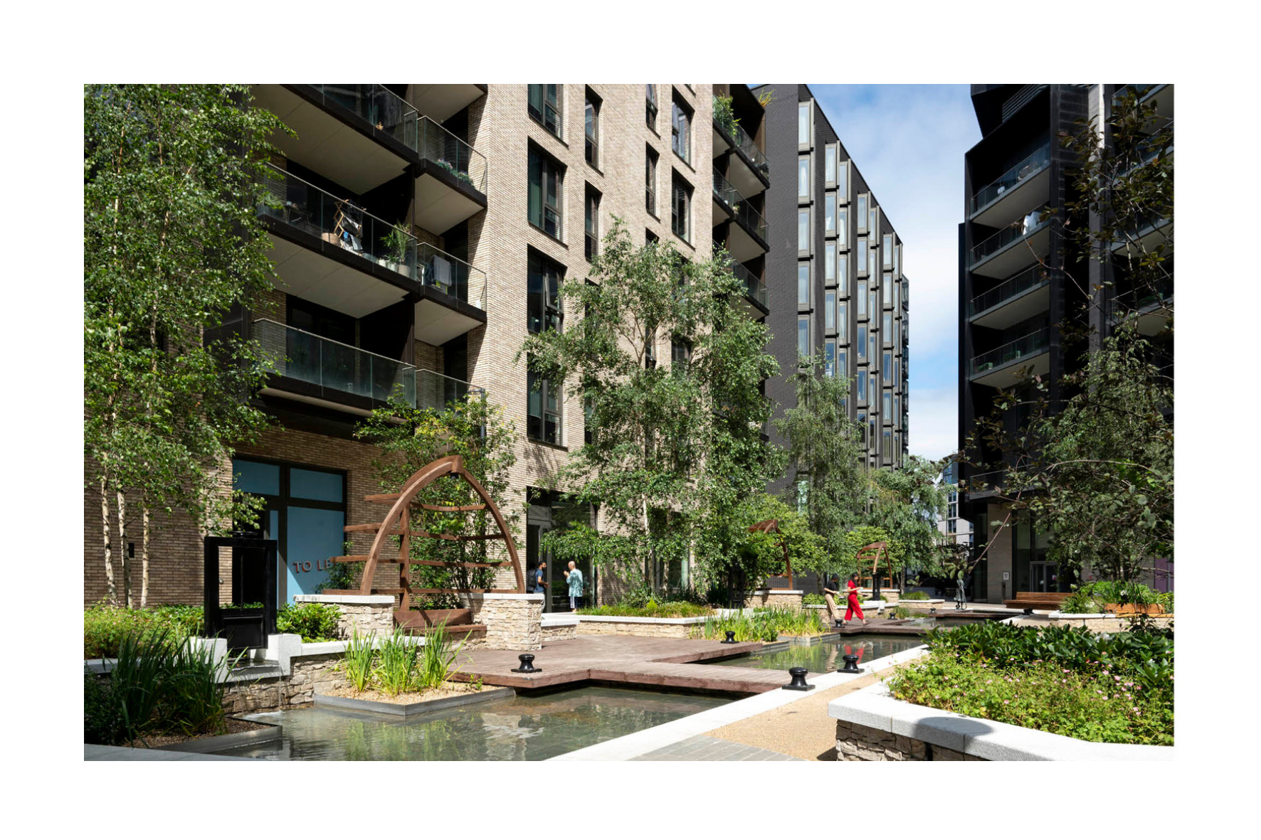 Modern residential apartment complex with landscaped courtyard, trees, benches, and a shallow water feature, with people walking and sitting.