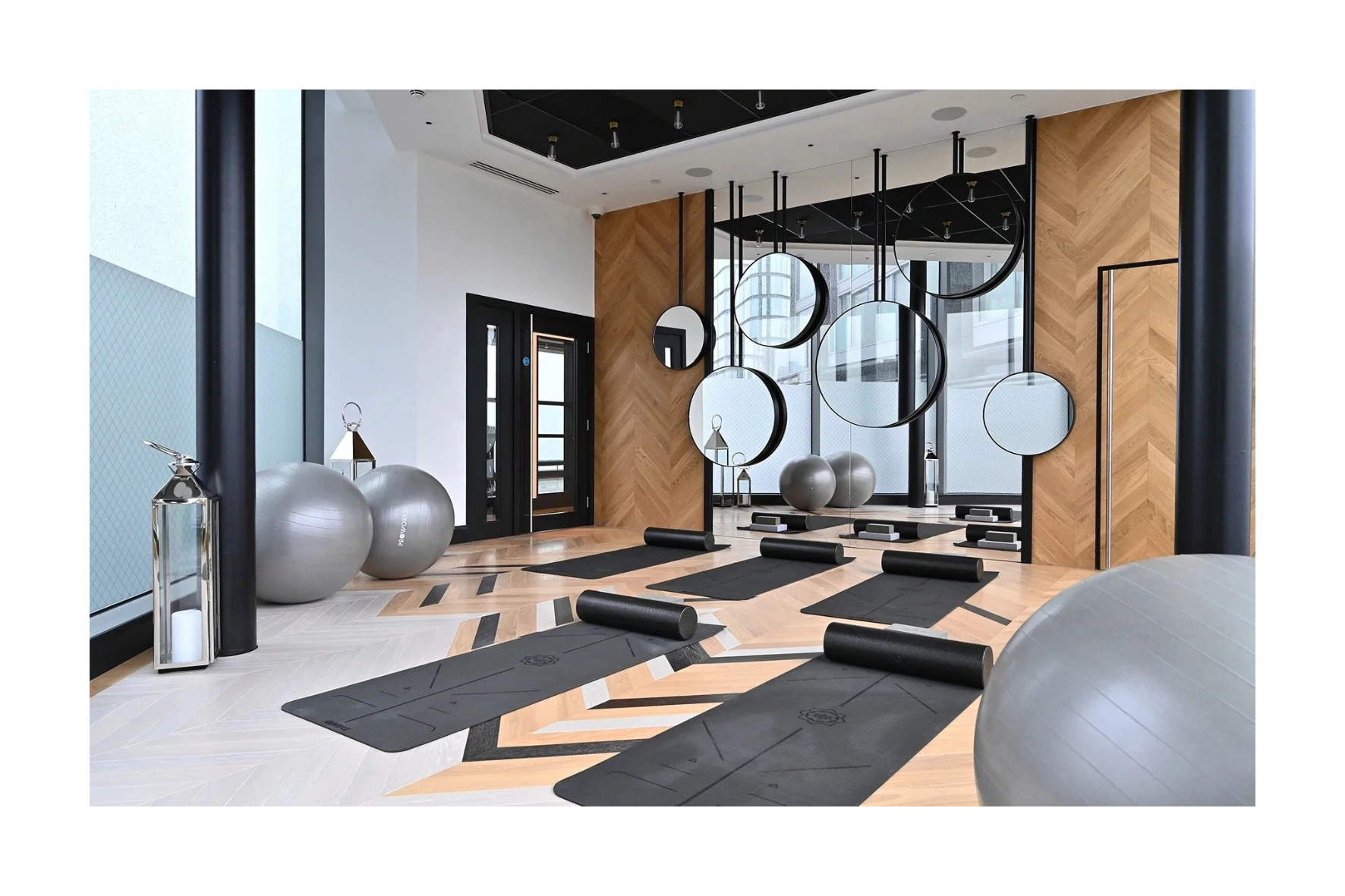 Indoor fitness studio with black yoga mats, foam rollers, and large exercise balls, decorated with round mirrors and wooden wall panels.