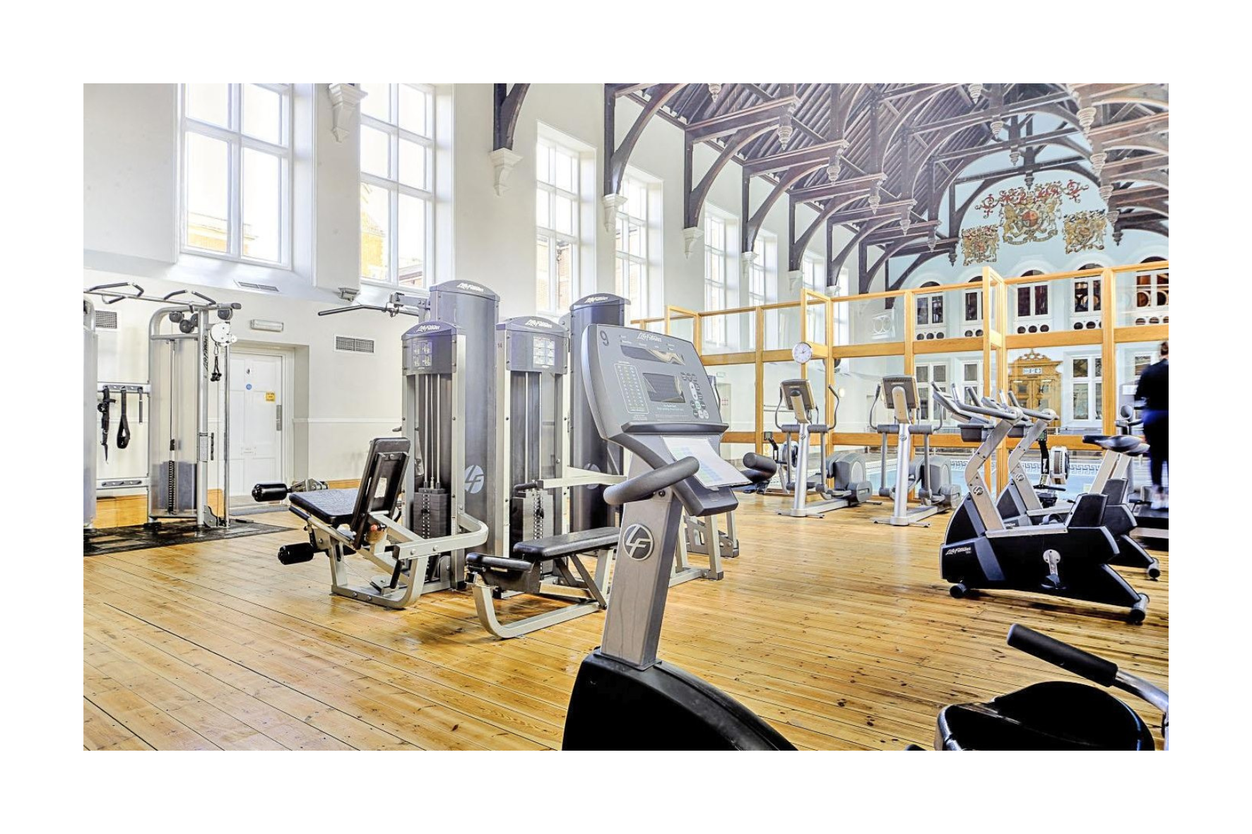 A spacious gym with high ceilings, large windows, and wooden floors, featuring various exercise equipment including treadmills, stationary bikes, and strength machines.