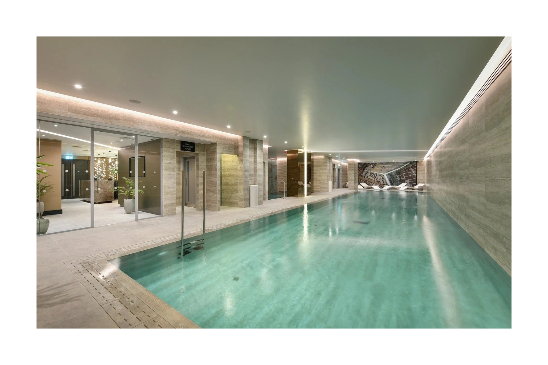 Indoor swimming pool area with lounge chairs along the wall at the far end, a tiled floor, and wall-mounted lights. There are glass doors leading to a changing room and a small pool or hot tub area.