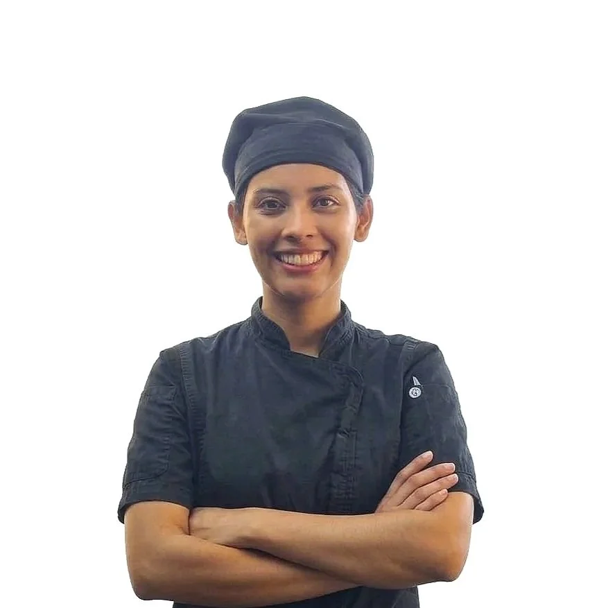 A smiling female chef with dark hair, wearing a black chef jacket and a black head wrap, standing with crossed arms against a plain white background.