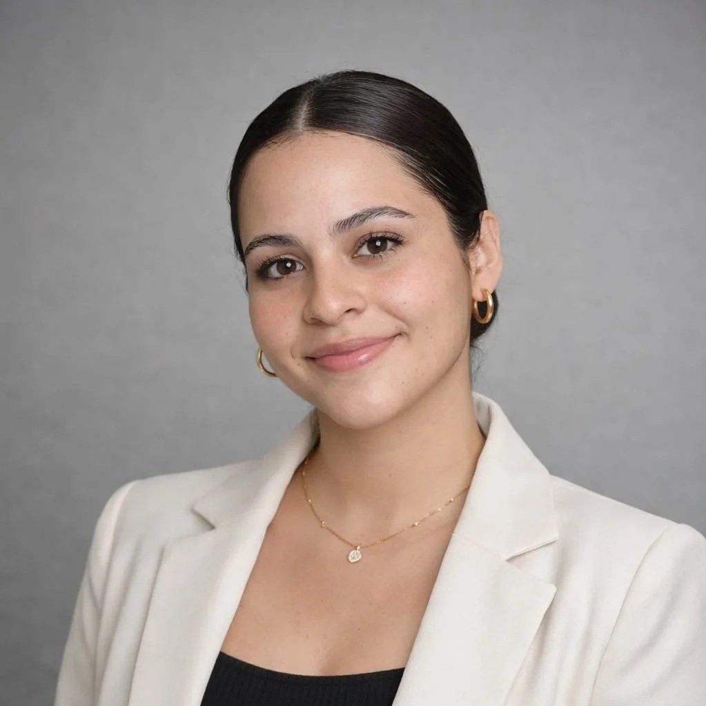 A woman with dark brown hair pulled back, wearing a white blazer, black top, gold hoop earrings, and a delicate gold necklace with a small pendant, smiling at the camera against a gray background.