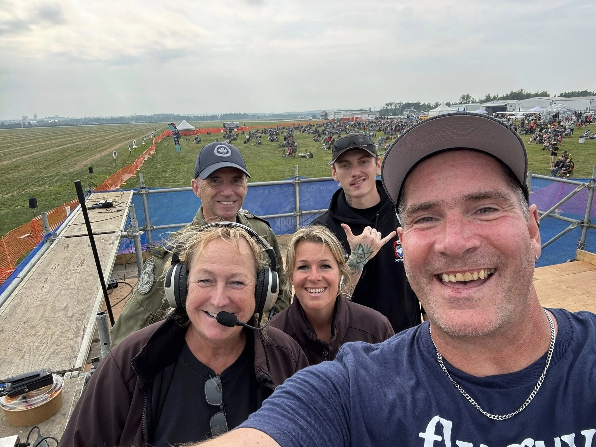 A group of five smiling people taking a selfie at an outdoor event with a large crowd and open field in the background.
