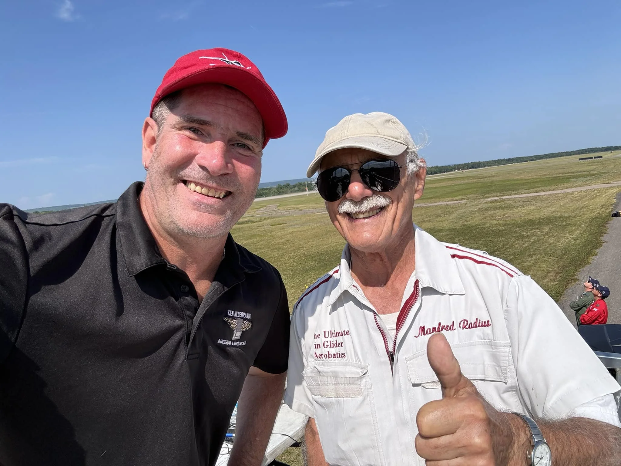 Two smiling men taking a selfie outdoors on a clear, sunny day. One man is wearing a black polo shirt and a red cap; the other is in a white shirt, sunglasses, and a beige cap, giving a thumbs-up. In the background, there is a grassy field and a pers