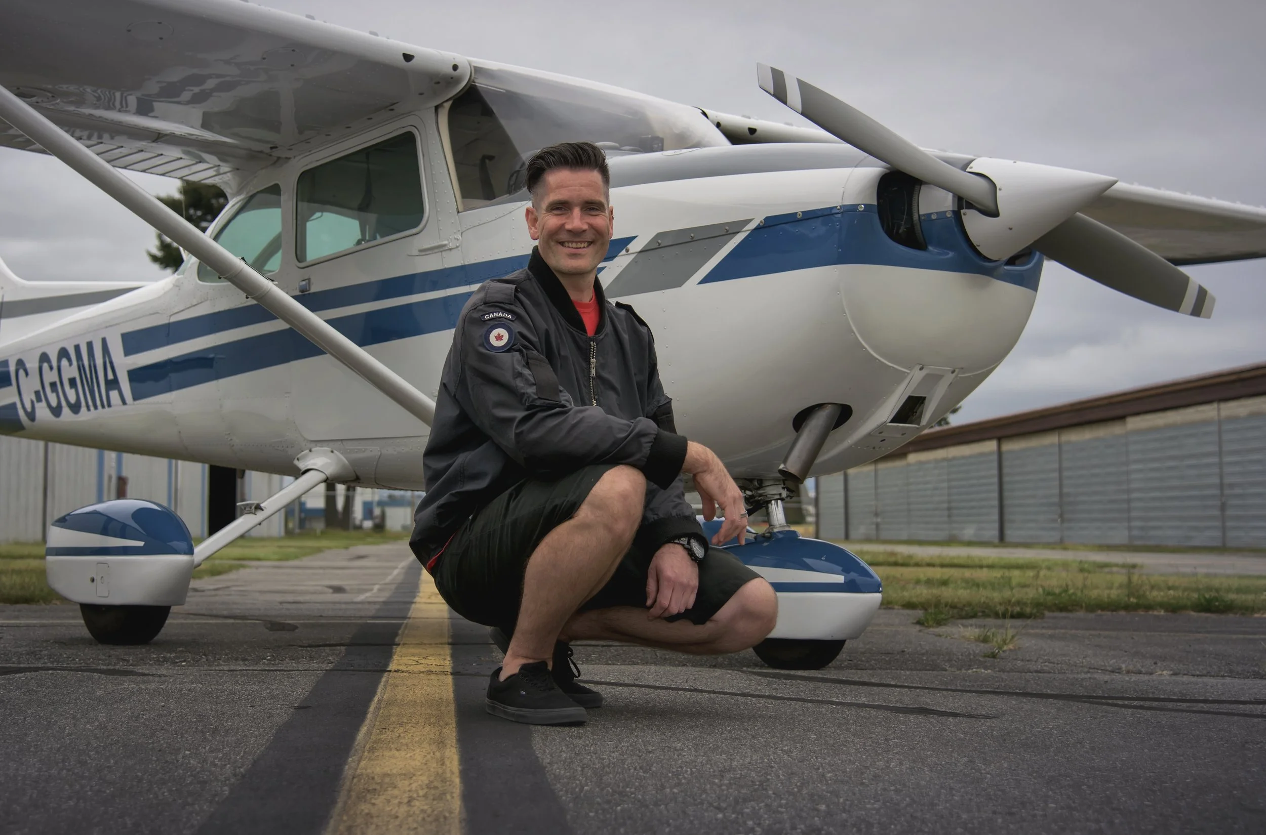 Man kneeling next to small white and blue propeller airplane on tarmac, smiling, wearing black jacket with Canada flag patch, and black shorts, with gray overcast sky in background.
