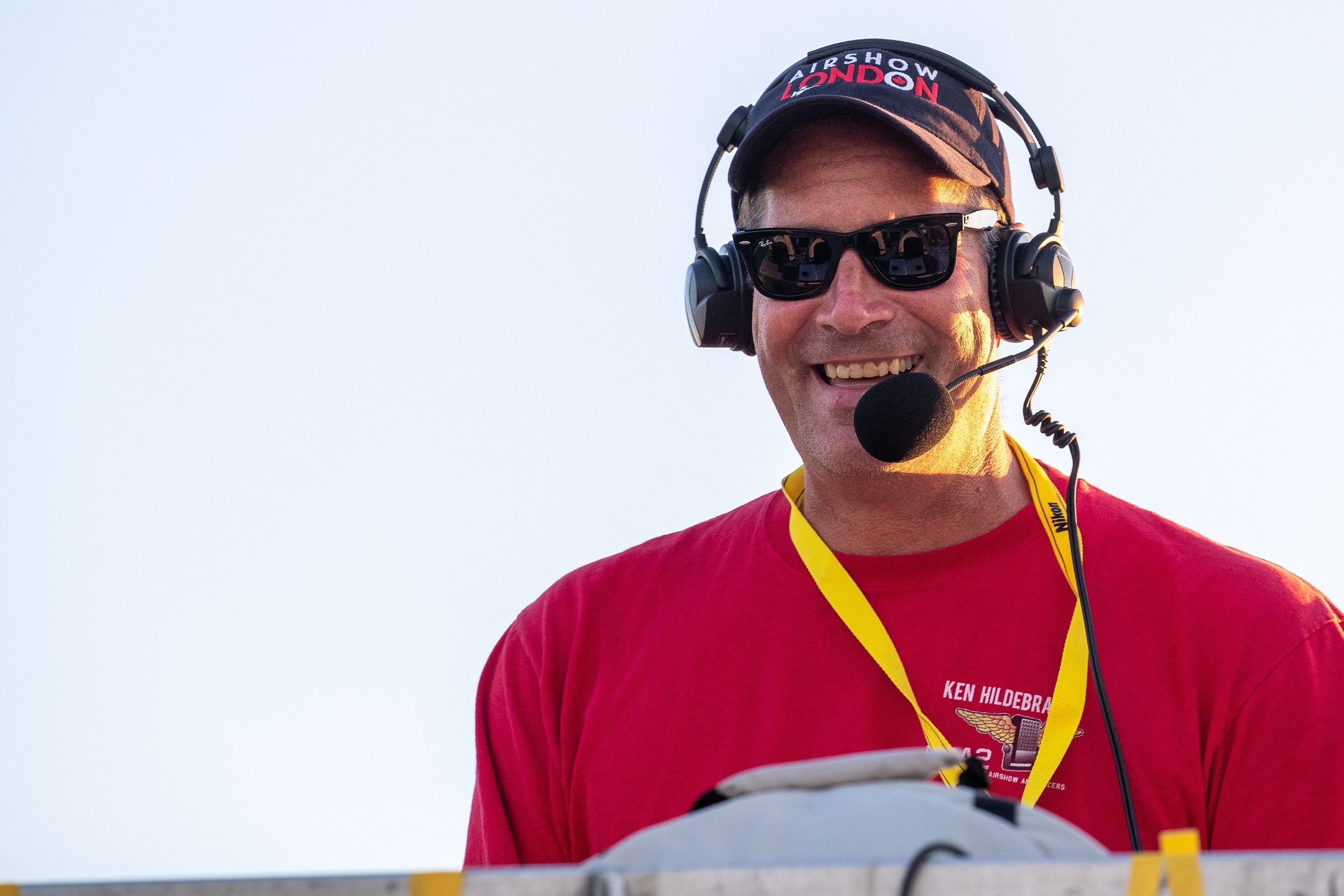 A smiling man wearing sunglasses, a headset with a microphone, a red T-shirt, and a cap that says 'Airshow London'.