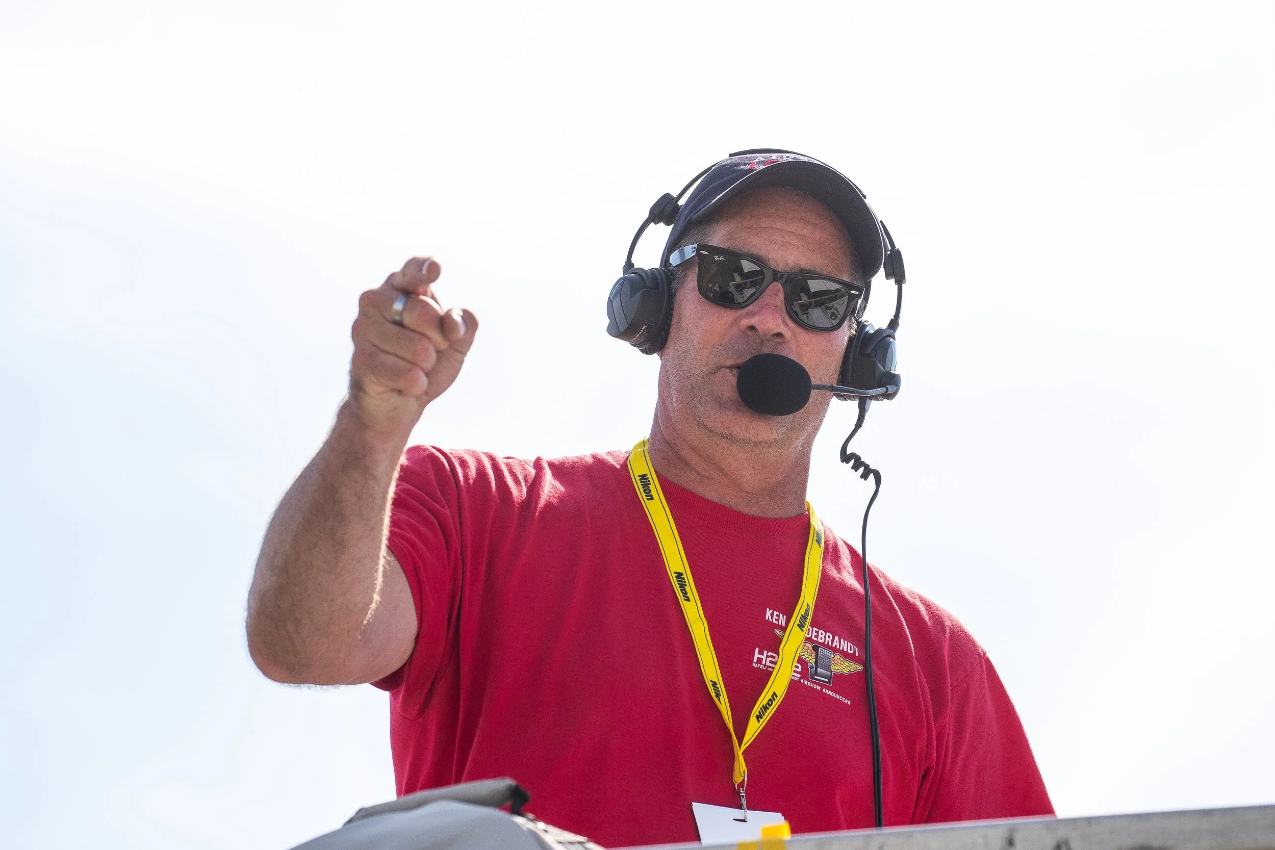 A man wearing sunglasses, headphones with a microphone, and a red shirt with multiple logos, gesturing with his hand during a broadcast or event.