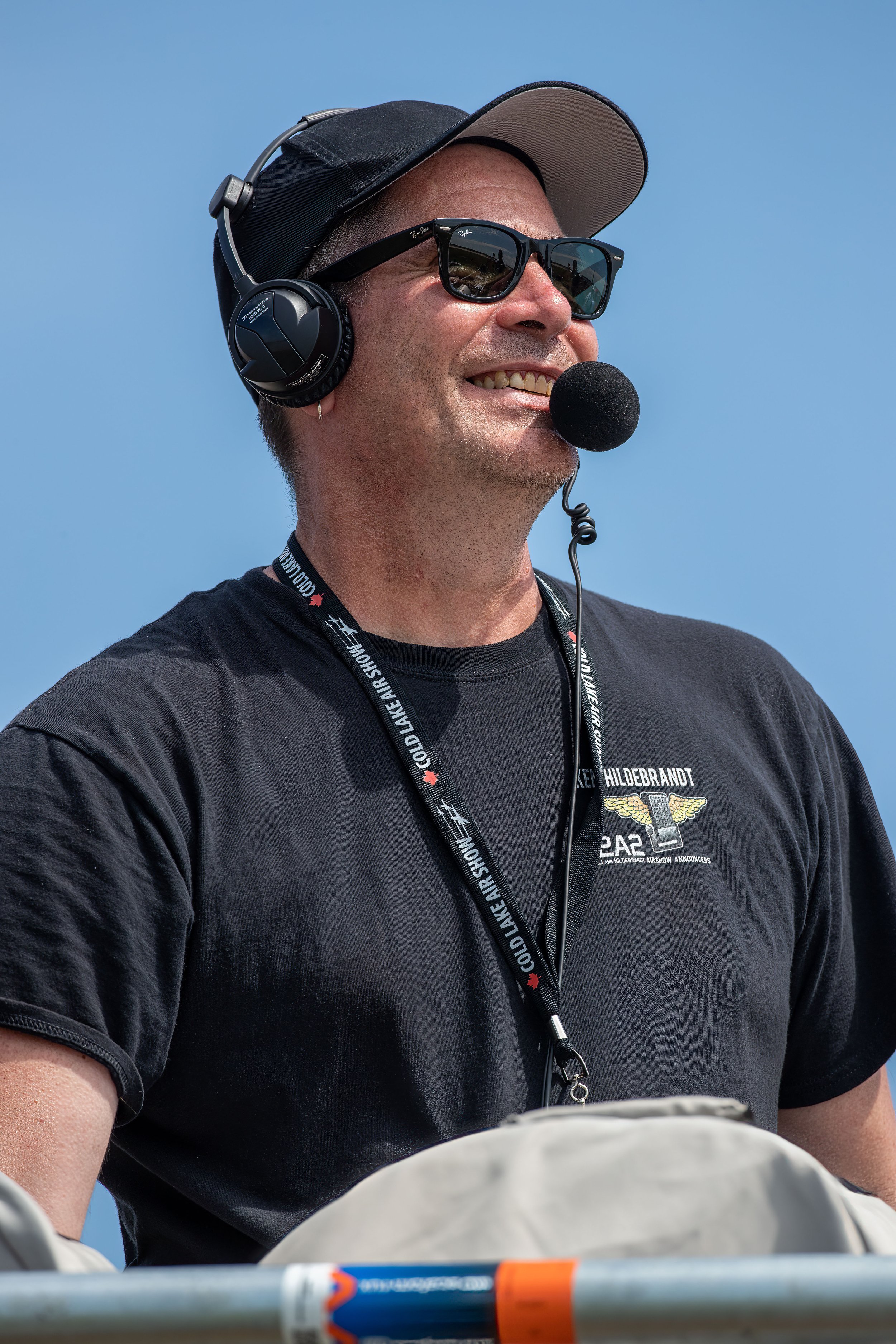 A man wearing a black T-shirt, sunglasses, hat, and headphones with a mic, smiling outdoors against a blue sky.