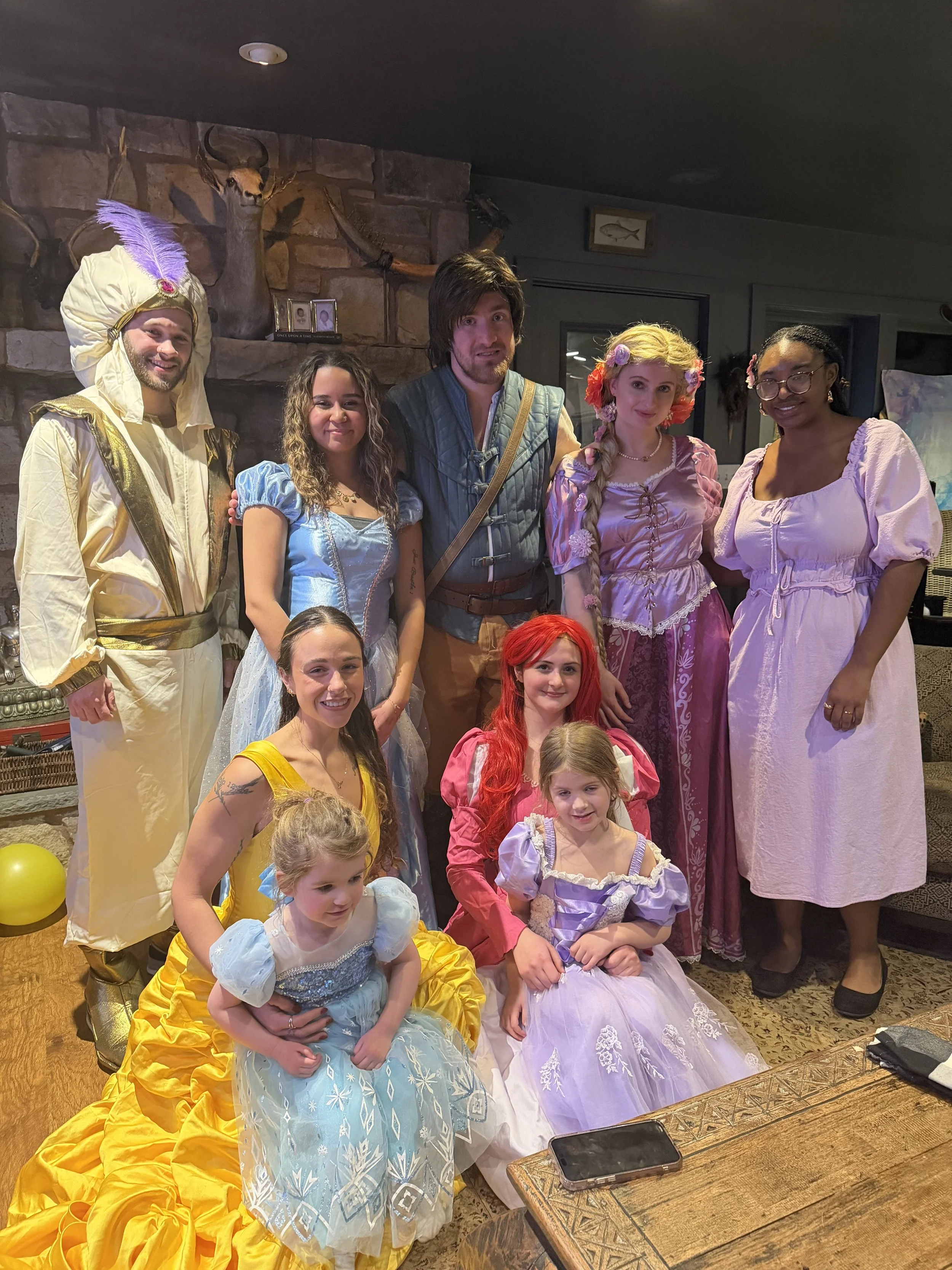 Group of people dressed in costumes inspired by fairy tale characters, including princesses and a man in a prince costume, posed indoors with a stone fireplace and mounted animal heads in the background.