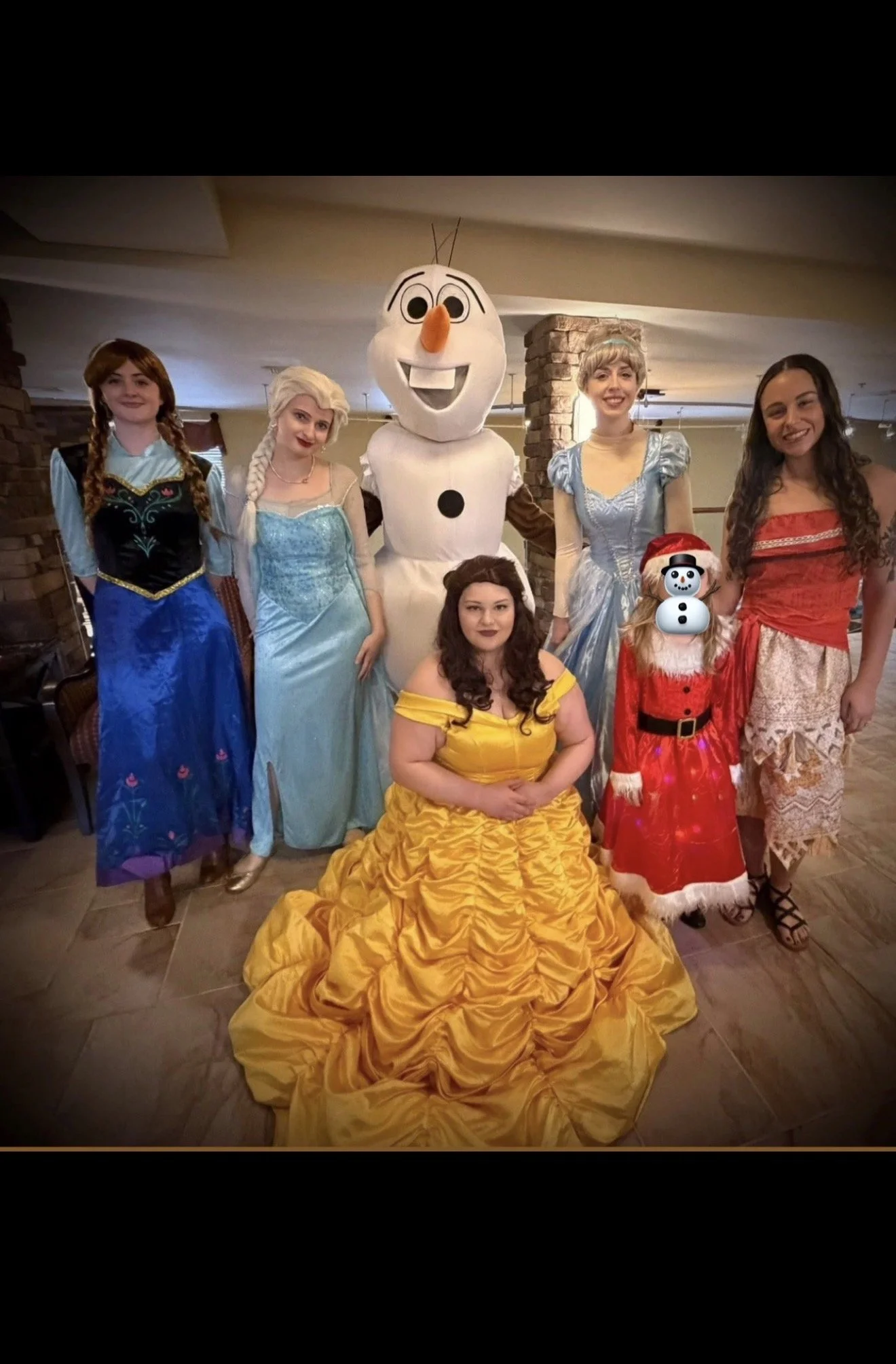 Group of five women dressed as Disney princesses and characters, including Belle, Elsa, Olaf, Snow Woman, and Anna, posing indoors with a woman in a yellow gown and a young girl dressed as Santa Claus.