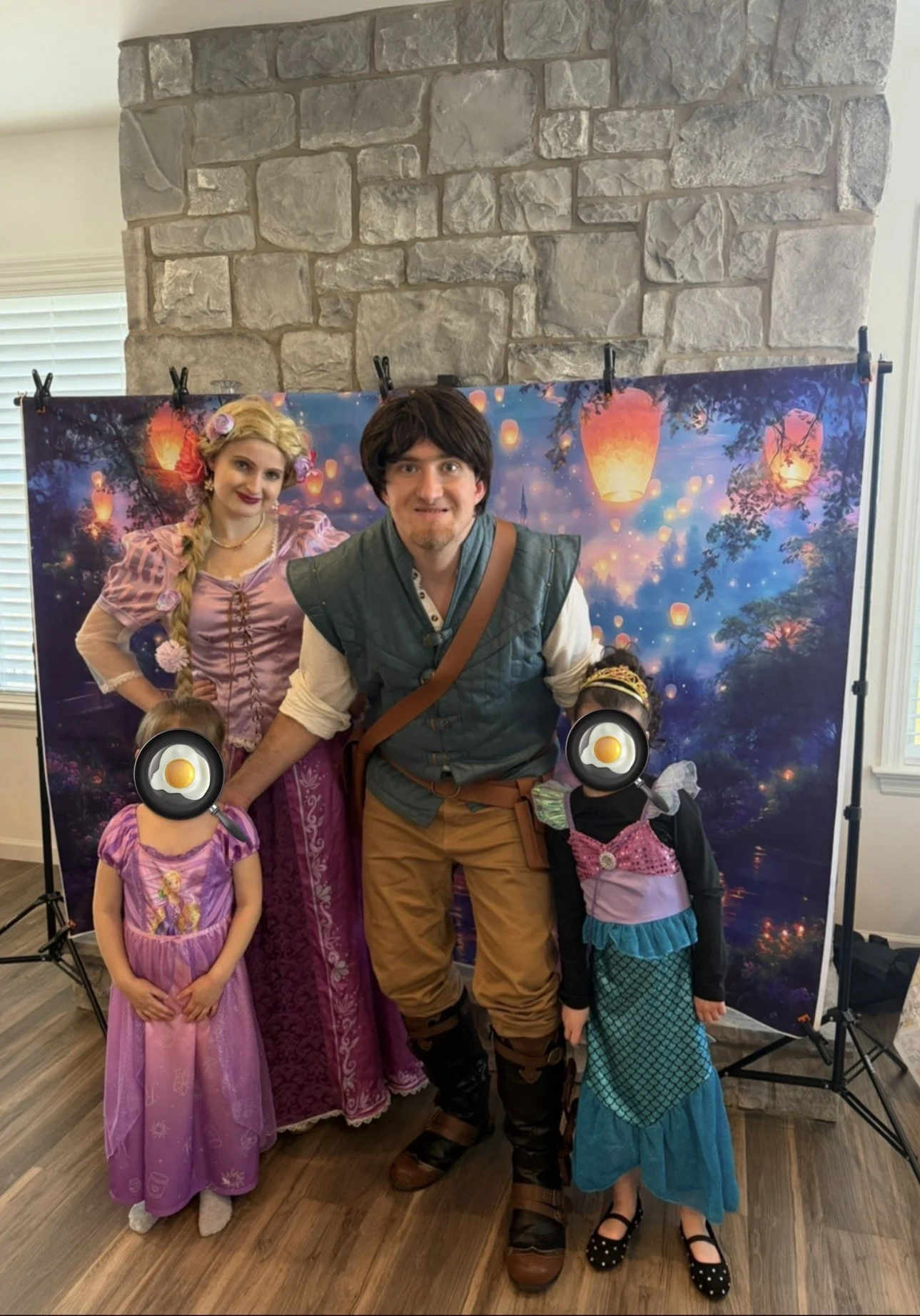 People dressed in costumes standing in front of a backdrop with floating lanterns at a lantern festival scene. Two children with faces obscured by fried egg cartoon stickers are in princess dresses. One adult man and one woman in princess dress are also present.