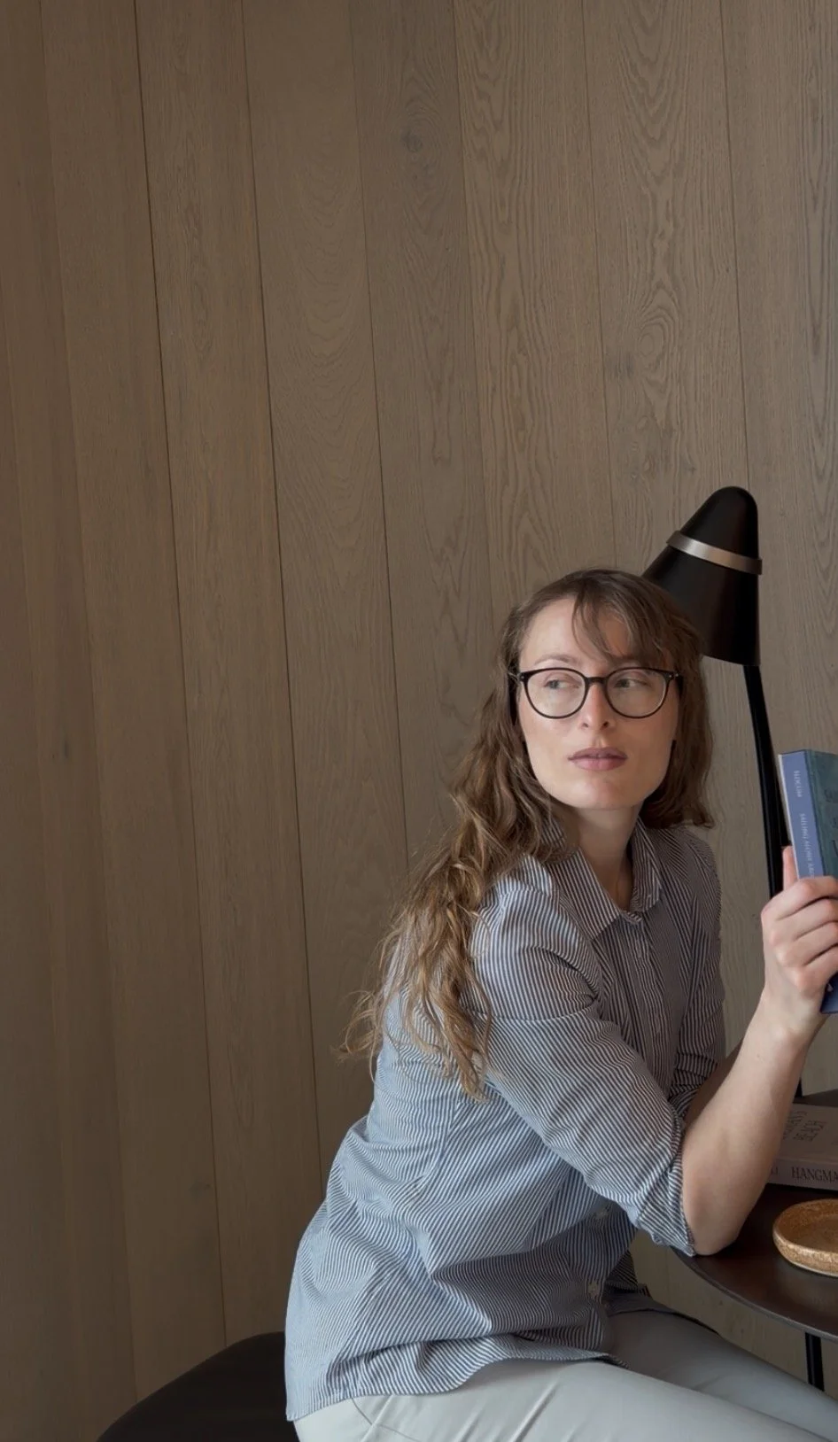 A woman with glasses and long wavy hair sitting at a table holding a book, against a wooden wall background.