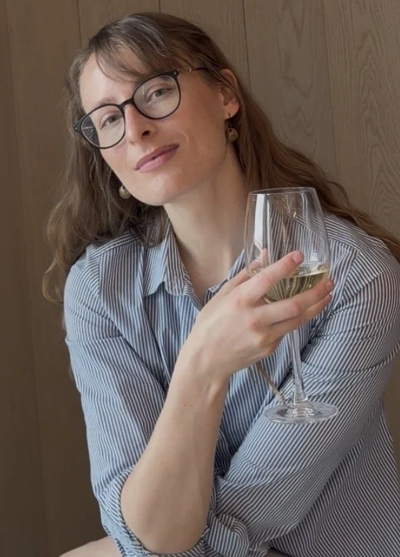 A woman with glasses and long hair holding a glass of white wine, sitting against a wooden wall.
